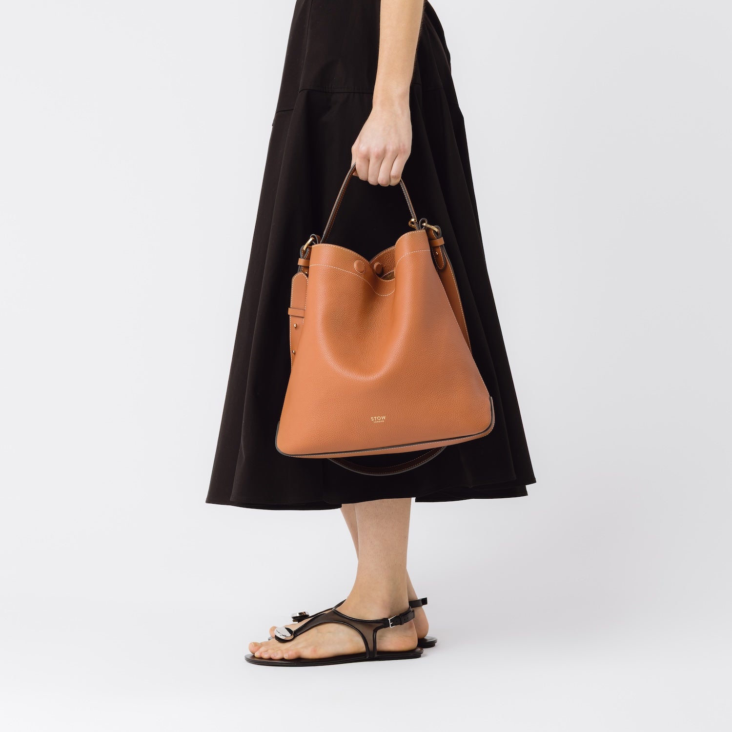 Model carrying STOW London's Curve bag in Earth Tan by its handle at her side featuring gold-coated stainless steel hardware on a white background