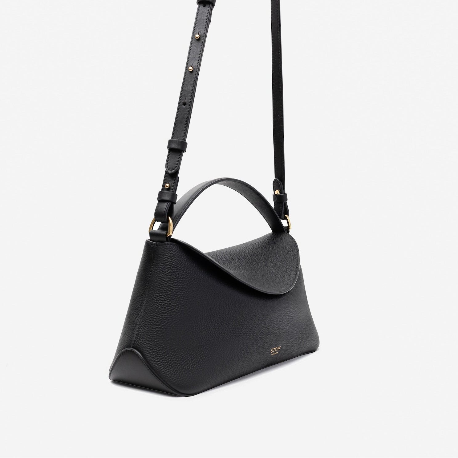Off-axis view of STOW London's East West in Black pebbled leather and detachable crossbody strap featuring gold-coated stainless steel hardware on a light grey background