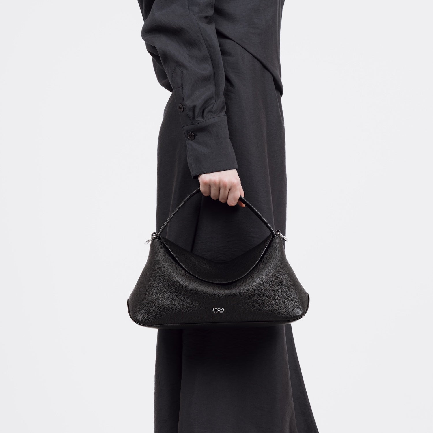 Close shot of model holding STOW London's East West bag in Black pebbled and smooth leather by its top handle at her side featuring silver stainless steel hardware on a white background