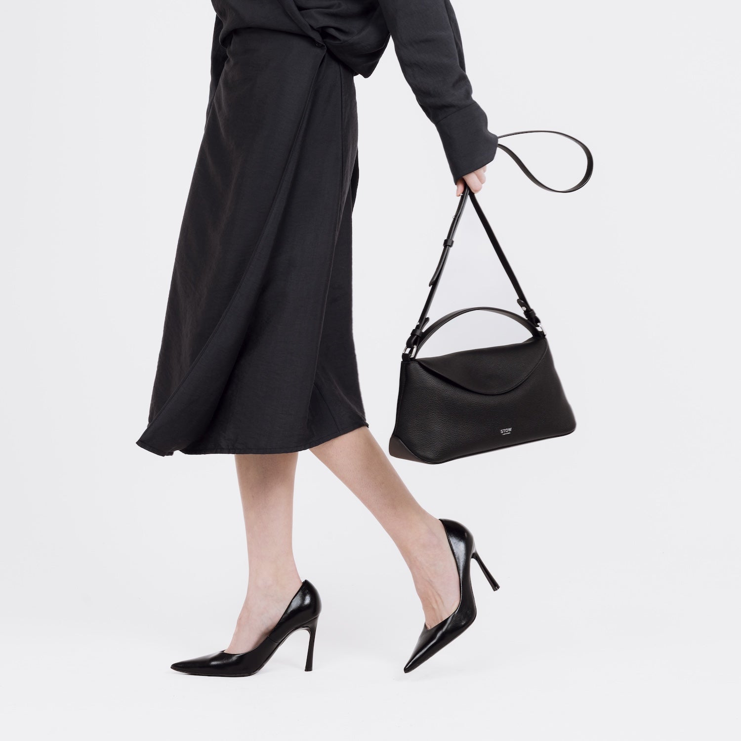 Model holding STOW London's East West bag in Black pebbled and smooth leather by its crossbody strap featuring silver stainless steel hardware on a white background
