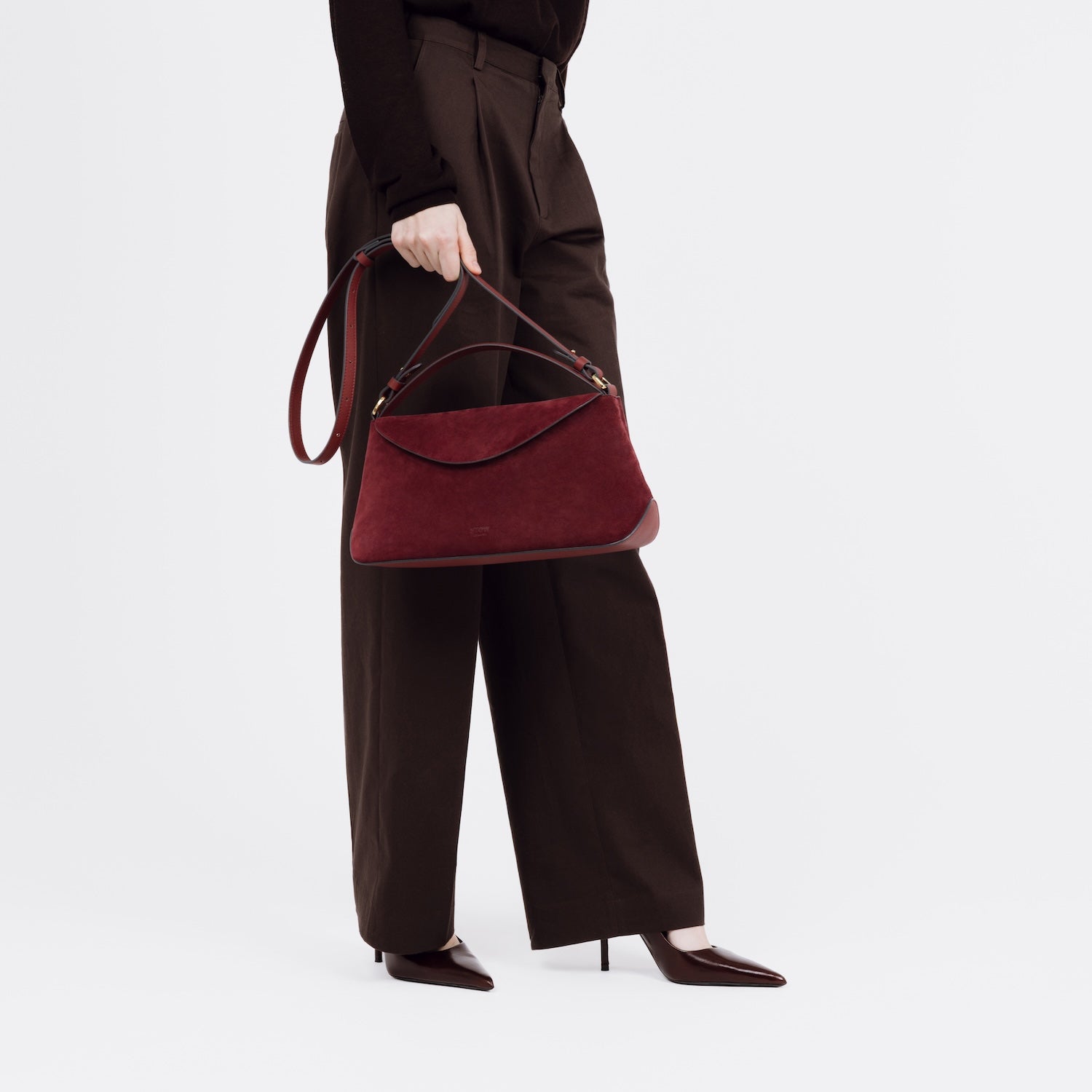Model holding STOW London's East West bag in Burgundy Suede leather by its detachable crossbody strap at her side featuring gold-coated stainless steel hardware on a white background