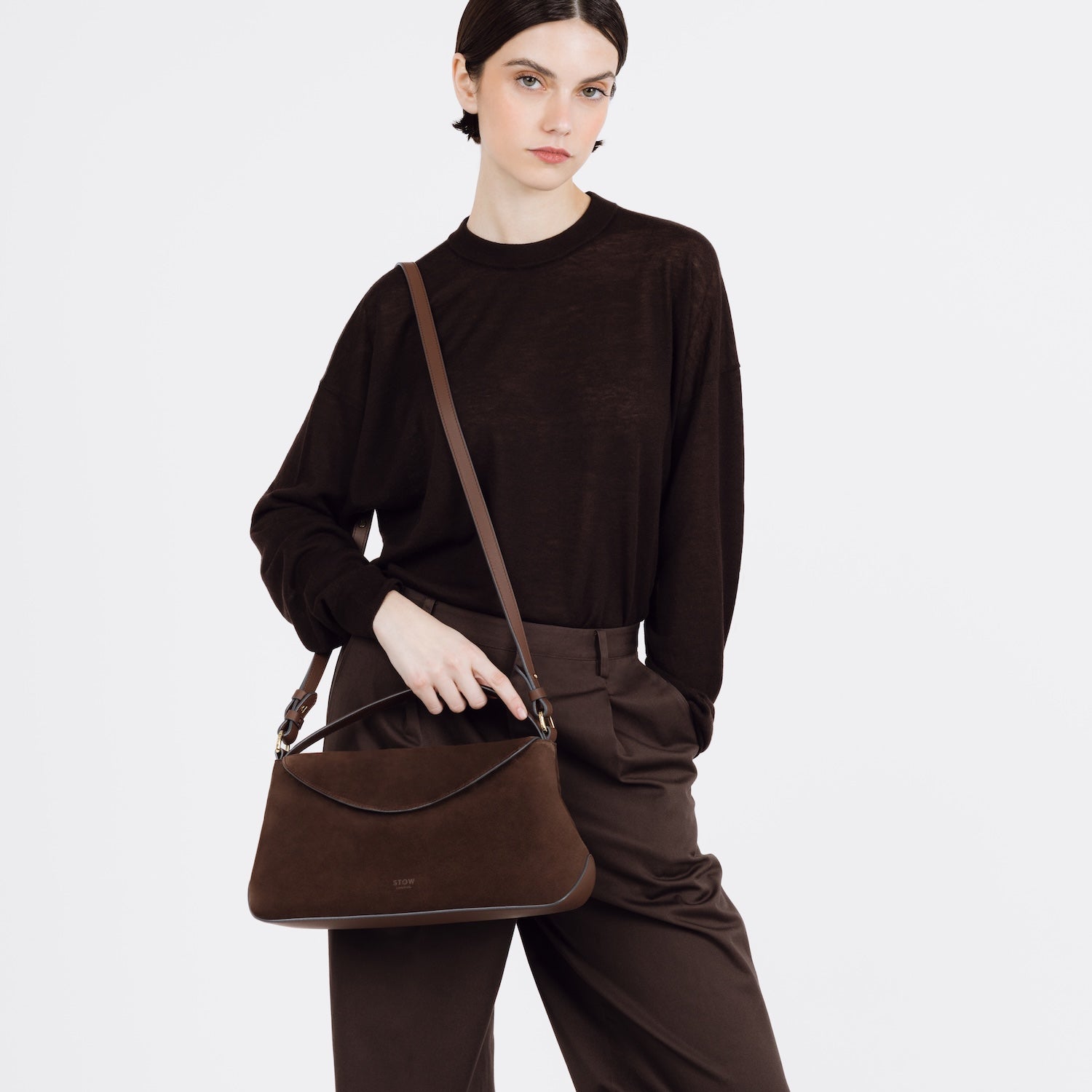 Model wearing STOW London's East West bag in Chocolate Suede leather on her shoulder featuring gold-coated stainless steel hardware on a white background
