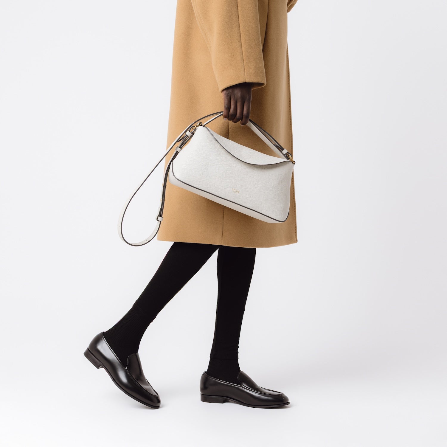 Model holding STOW London's East West in Cloud Grey pebbled leather at her side featuring gold-coated stainless steel hardware on a light grey background