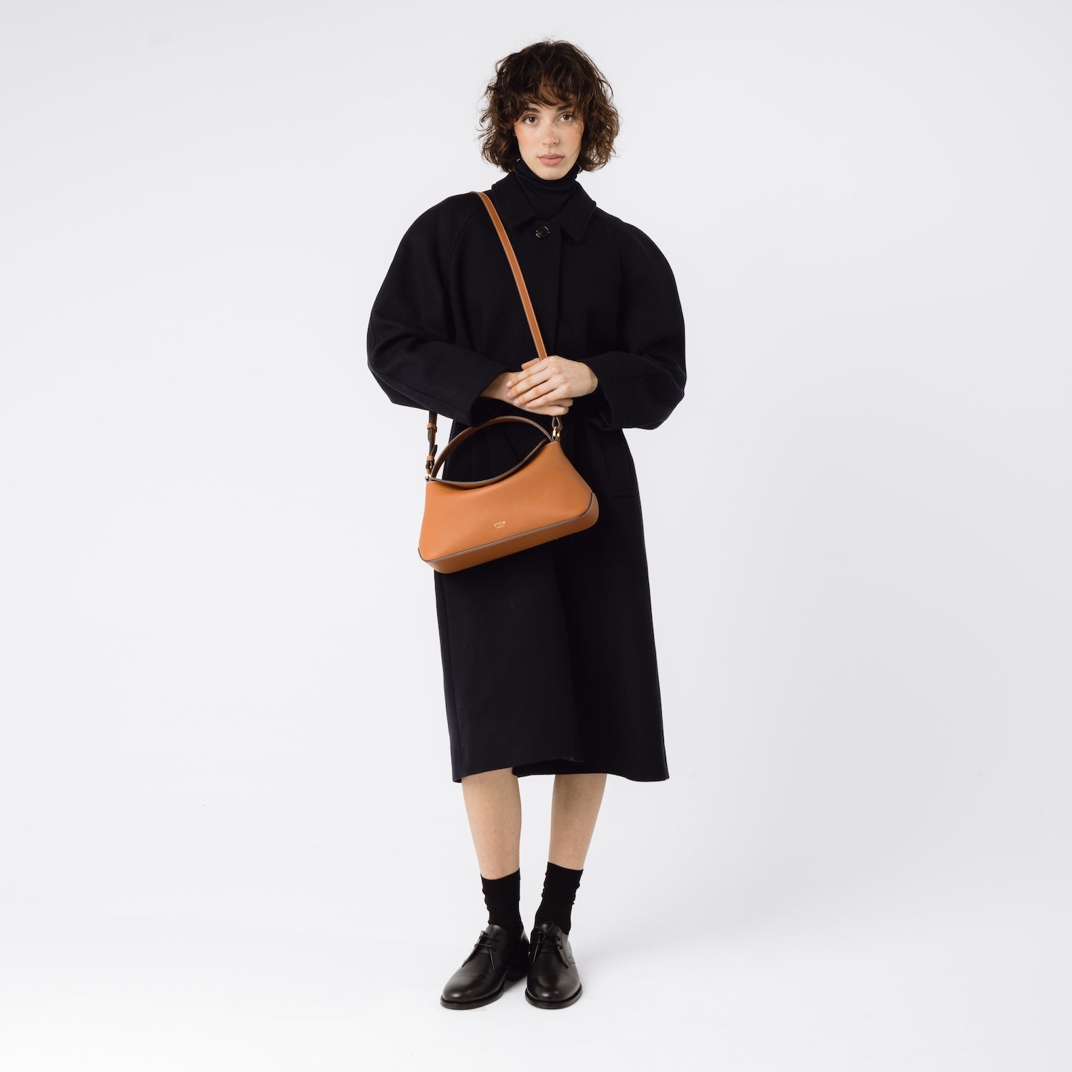 STOW London's East West bag in Earth Tan featuring gold-coated stainless steel hardware worn over model's shoulder by detachable strap on a white background