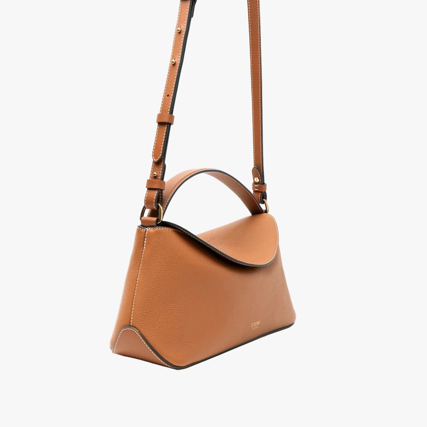 Off-axis view of STOW London's East West bag in Earth Tan pebbled leather and crossbody strap featuring gold-coated stainless steel hardware on a light grey background