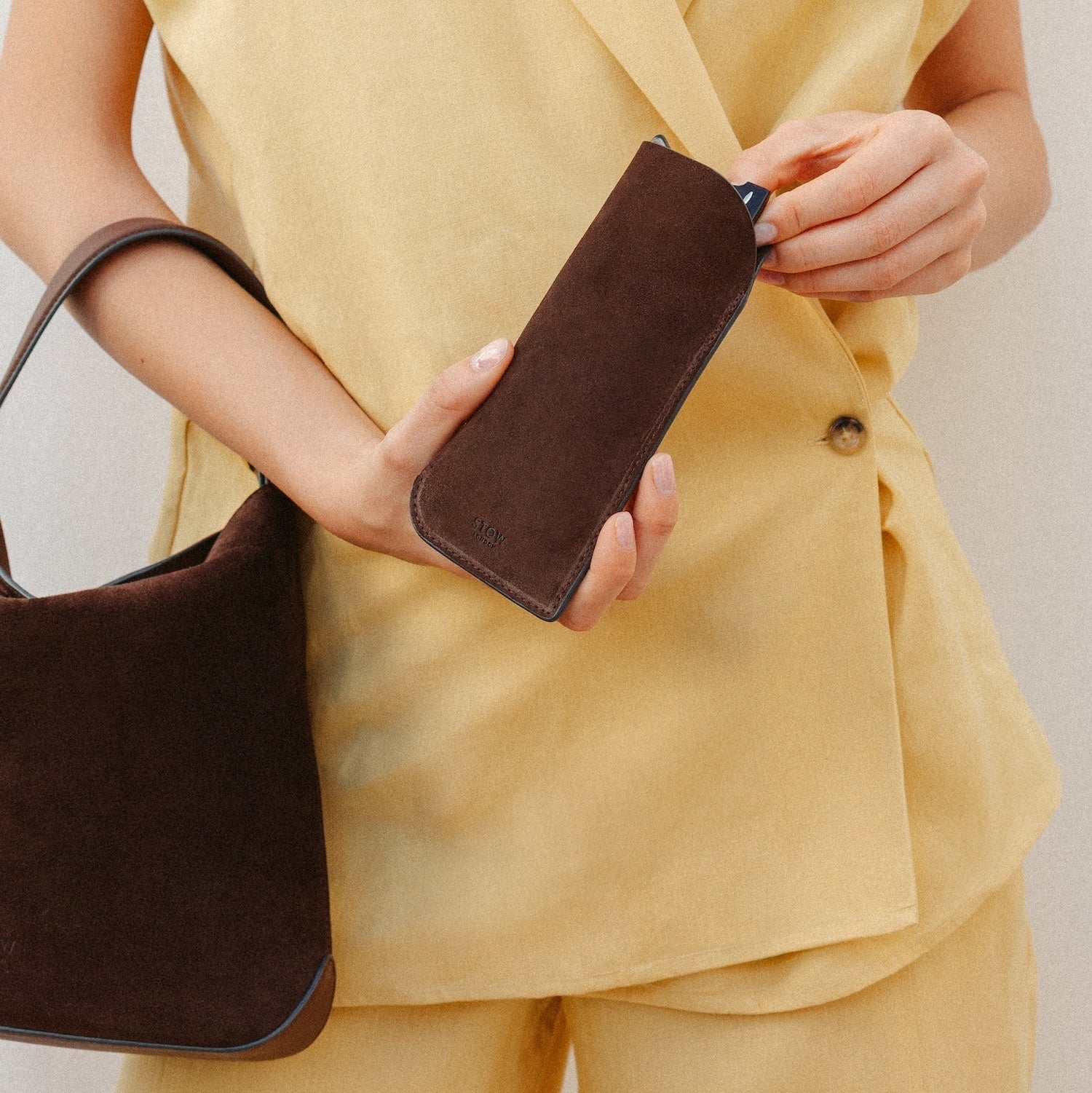Model putting her glasses into STOW London's Glasses Case in Chocolate suede leather with the Chocolate suede Midi Curve on her arm