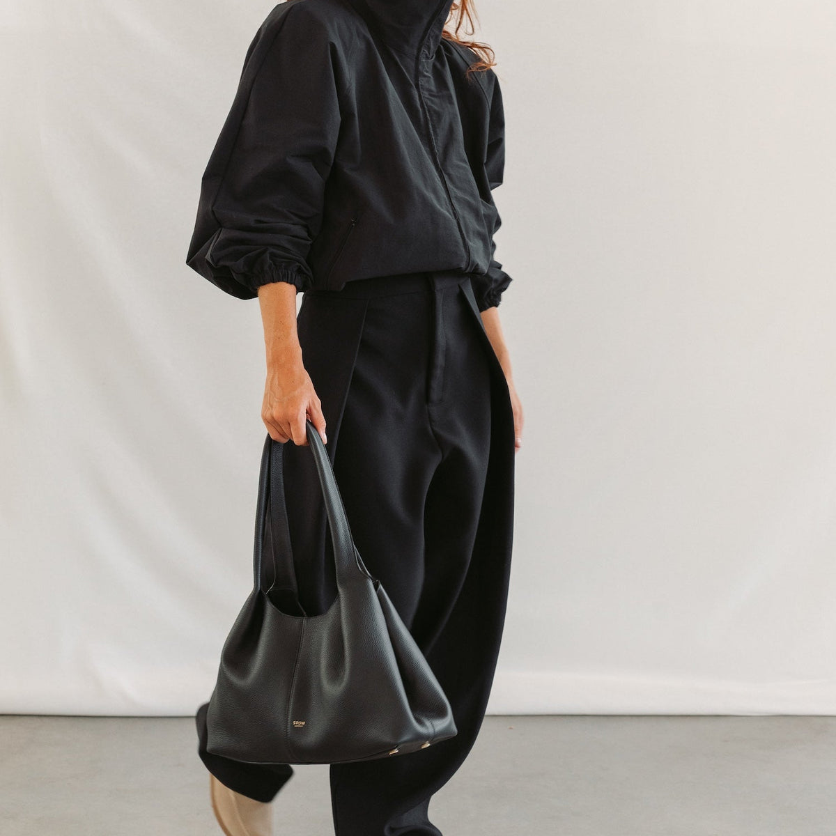 Model holding STOW London's Hobo Tote in Black pebbled leather featuring a pleated front panel and gold-coated stainless steel hardware
