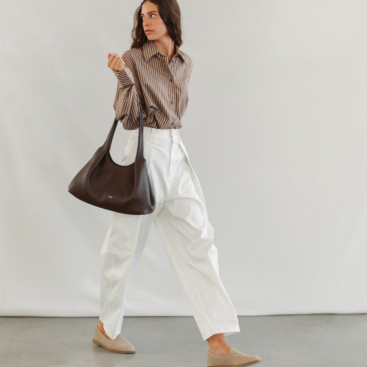 Model wearing STOW London's Hobo Tote in Espresso pebbled leather featuring a pleated front panel and gold-coated stainless steel hardware