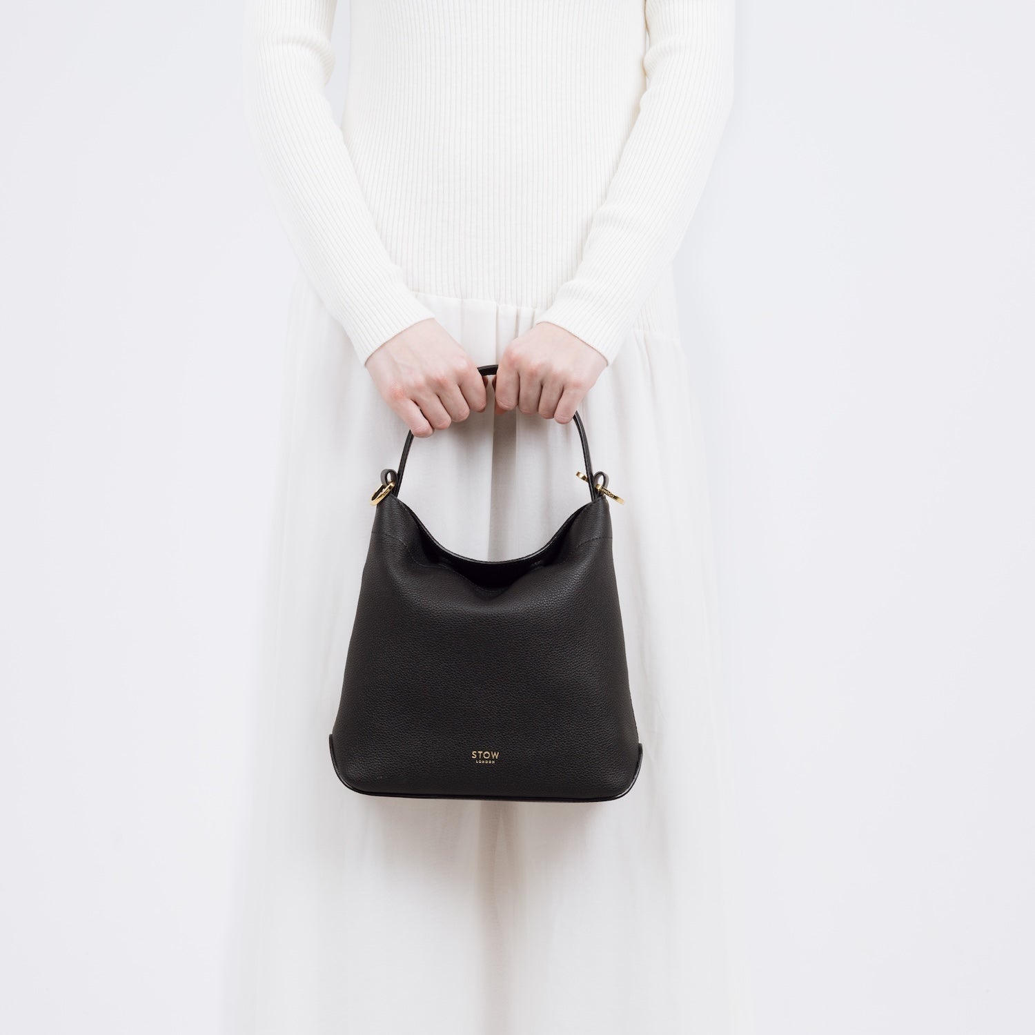Model holding STOW London's Midi Curve bag in Black pebbled and smooth leather at her front in a classic flap bag style featuring gold-coated stainless steel hardware on a white background