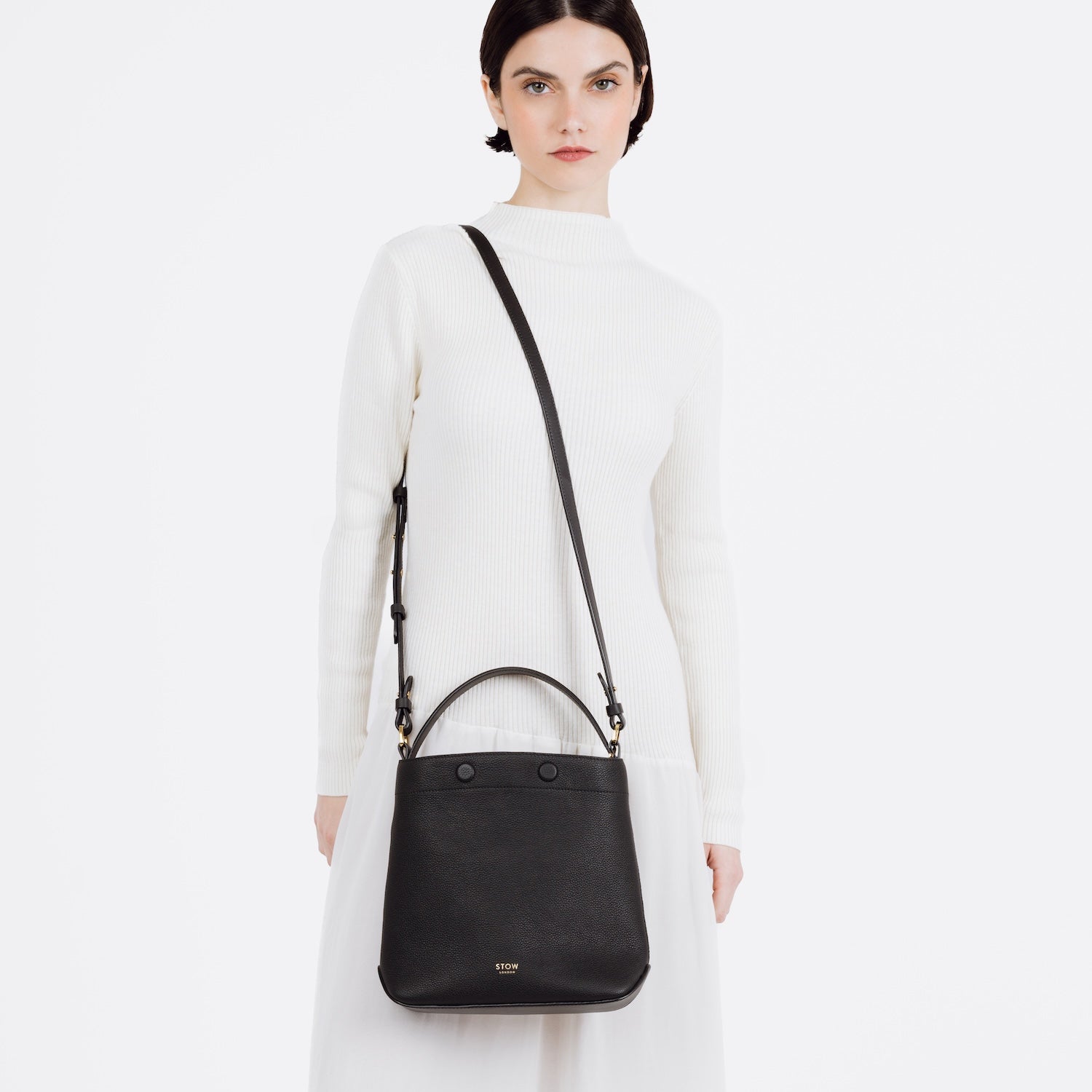 Model wearing STOW London's Midi Curve bag in Black pebbled and smooth leather over her shoulder in a bucket bag style featuring gold-coated stainless steel hardware on a white background