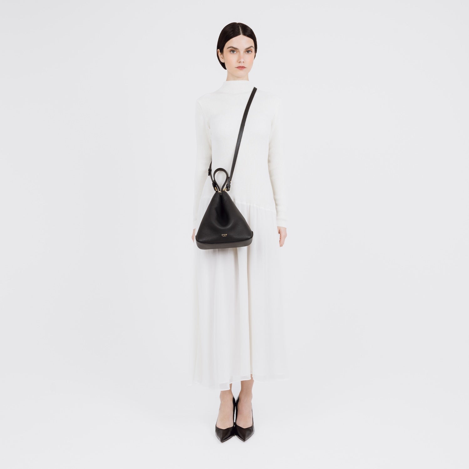 Model wearing STOW London's Midi Curve bag in Black pebbled and smooth leather crossbody in a triangular style featuring gold-coated stainless steel hardware on a white background