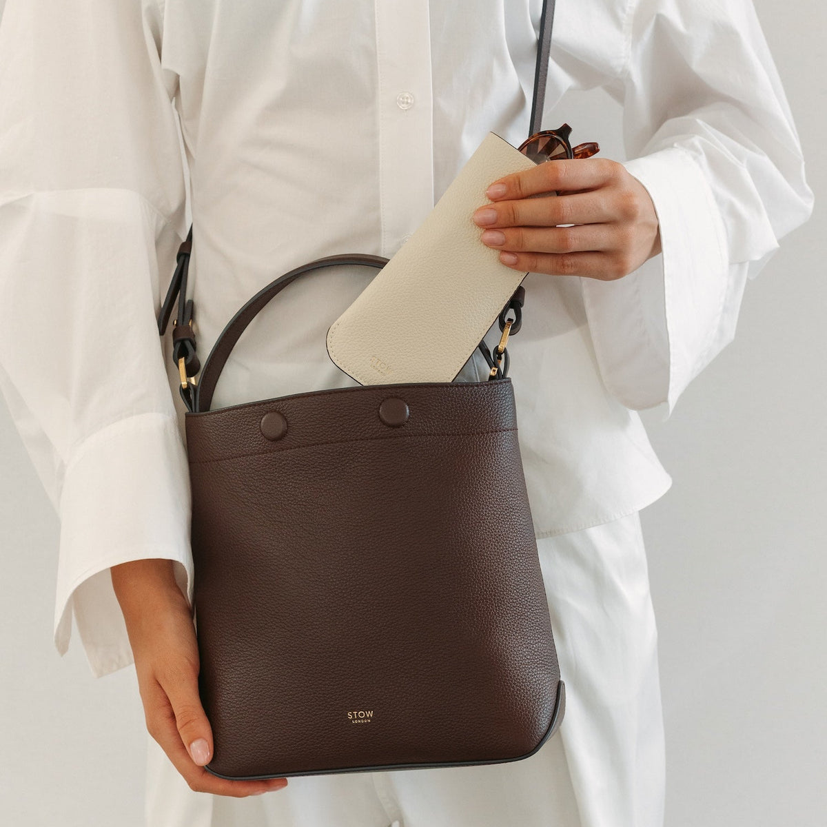 Model in a white outfit wearing STOW London's Midi Curve in Espresso pebbled leather featuring the Spring Moon Glasses case and gold-coated stainless steel hardware