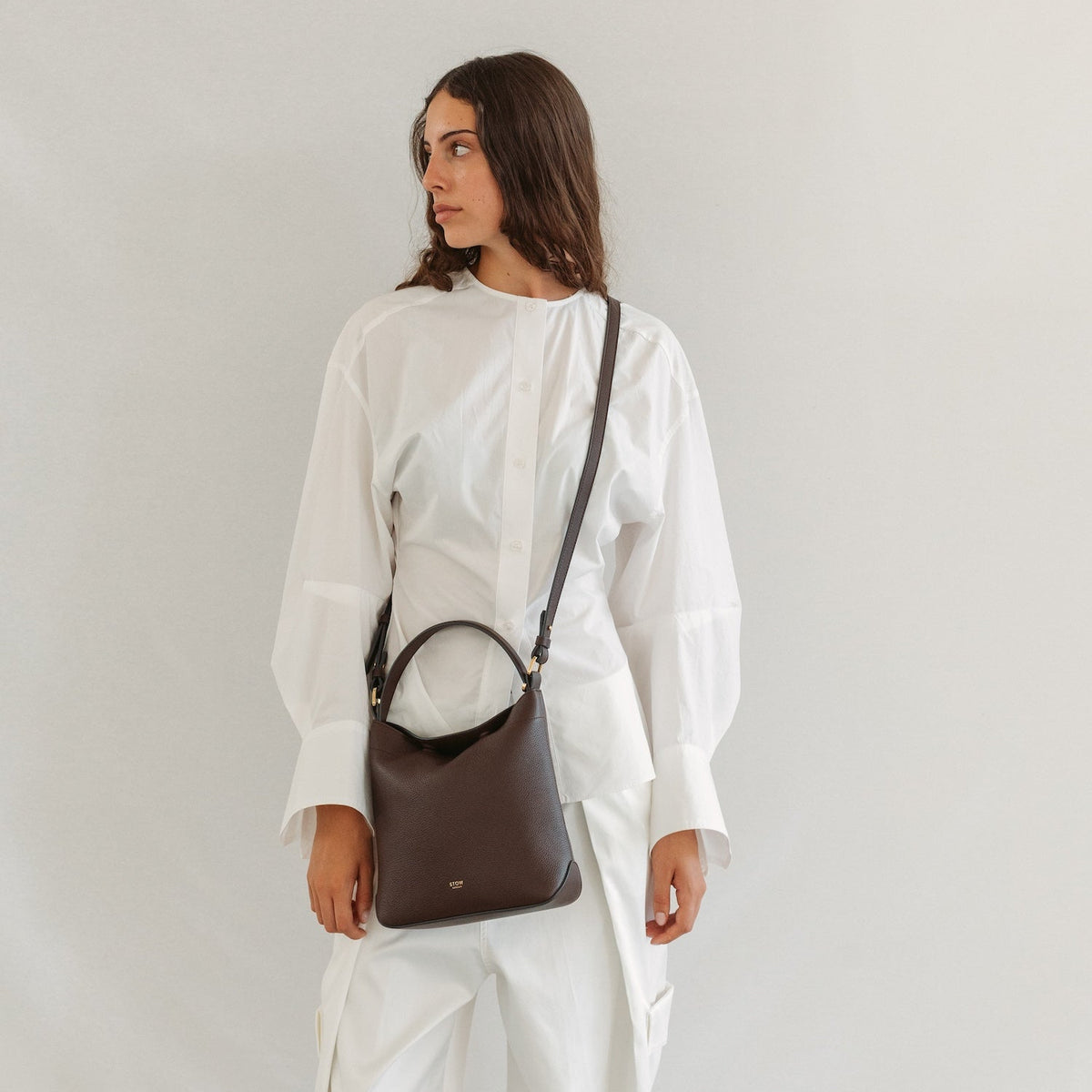 Model in a white outfit wearing STOW London's Midi Curve in Espresso pebbled leather crossbody featuring gold-coated stainless steel hardware