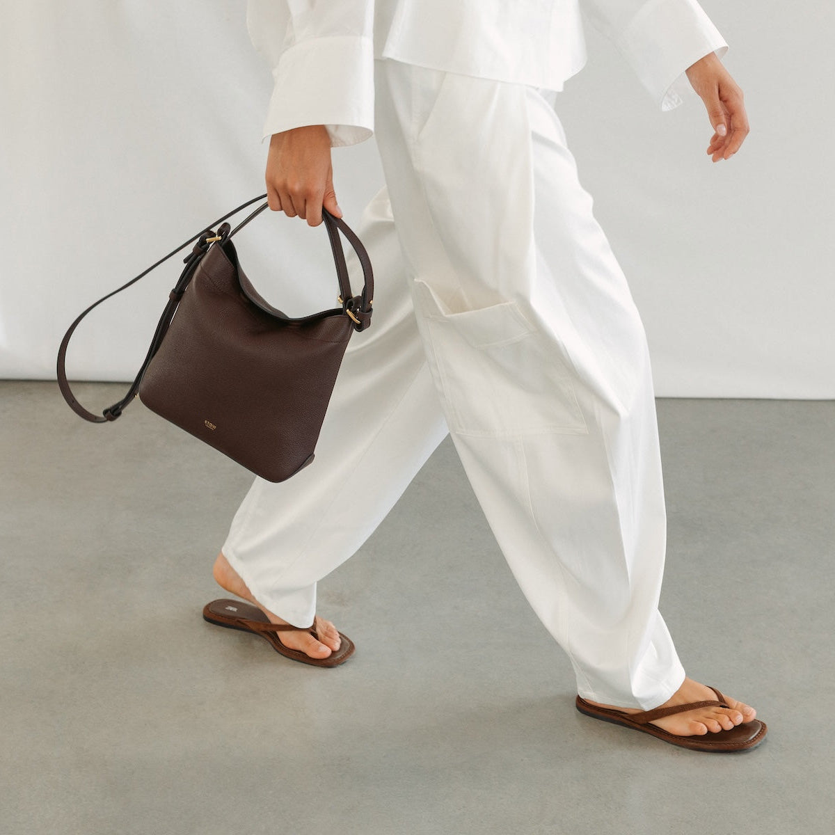 Model in a white outfit holding STOW London's Midi Curve in Espresso pebbled leather featuring gold-coated stainless steel hardware