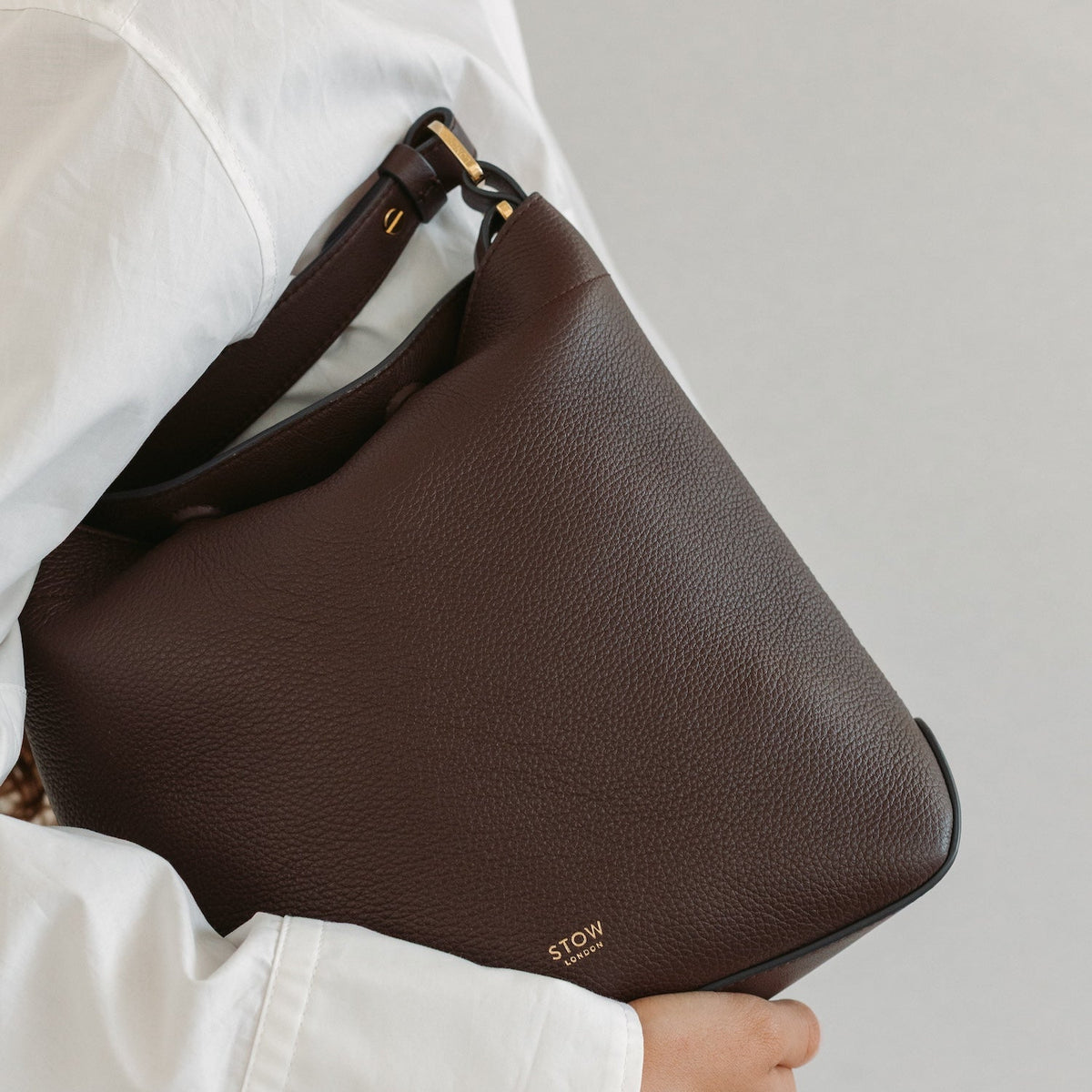 Model in a white outfit holding STOW London's Midi Curve in Espresso pebbled leather featuring gold-coated stainless steel hardware and small embossed logo