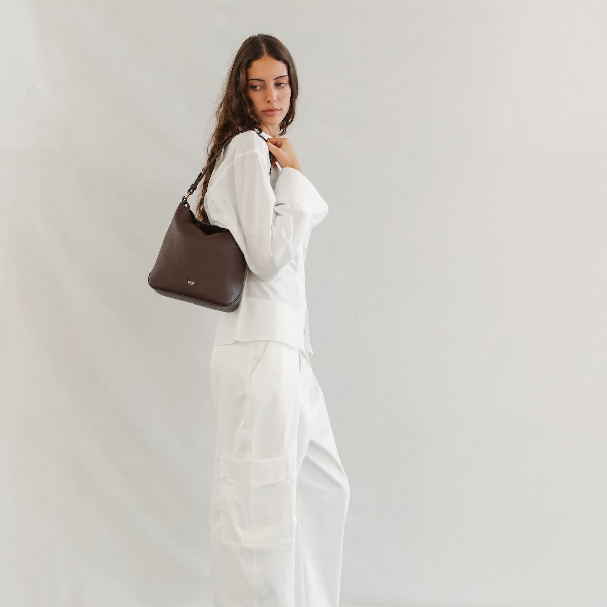 Model wearing STOW London's Midi Curve in Espresso pebbled leather featuring the detachable shoulder strap and gold-coated stainless steel hardware
