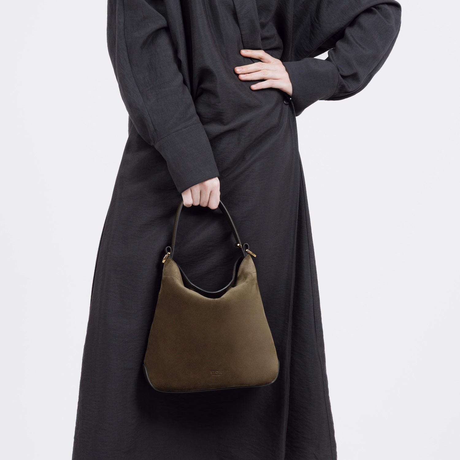 Model holding STOW London's Midi Curve bag in Dark Green Suede leather by the top handle at her front styled as a flap bag featuring gold-coated stainless steel hardware on a white background