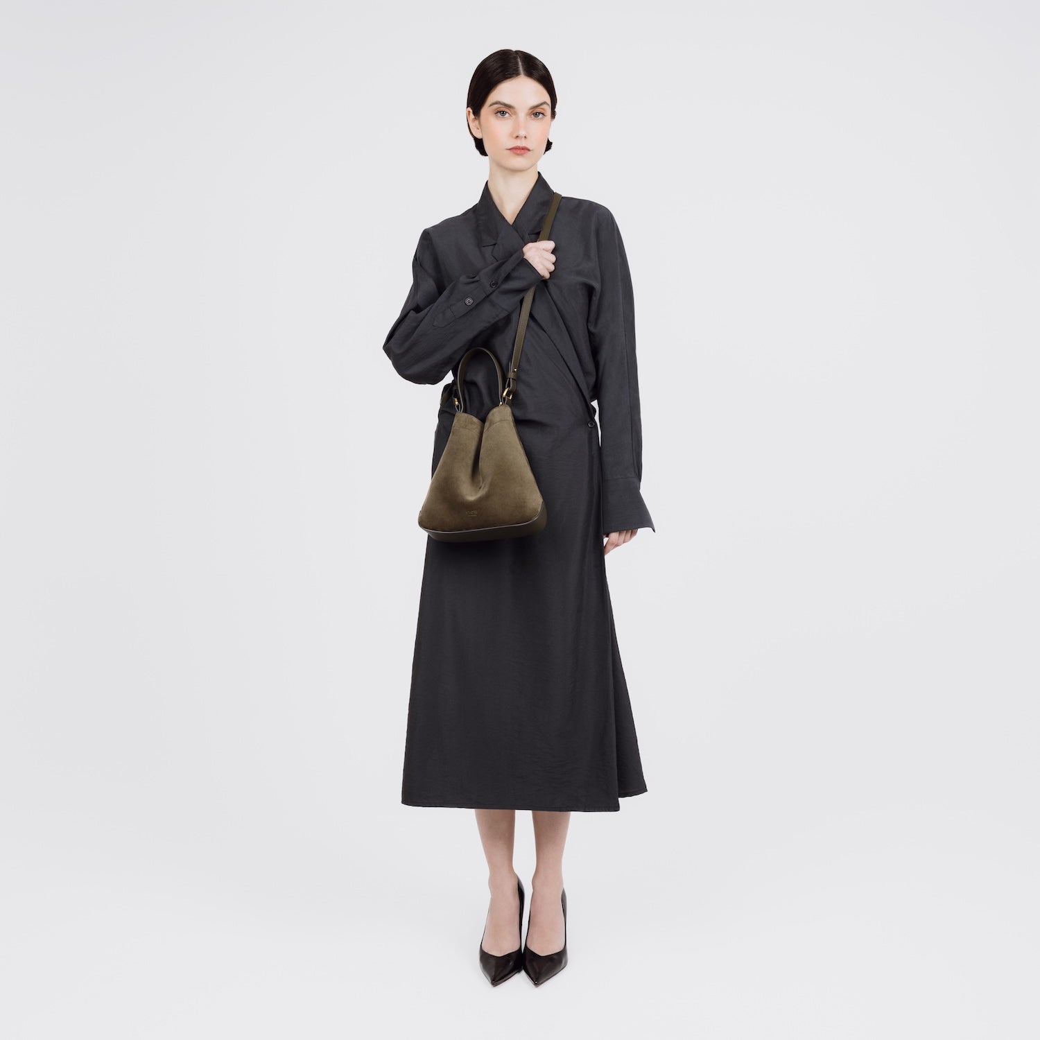 Model wearing STOW London's Midi Curve bag in Dark Green Suede leather crossbody styled in a triangular shape featuring gold-coated stainless steel hardware on a white background