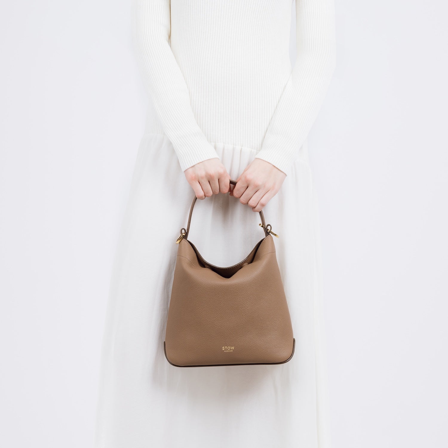 Model holding STOW London's Midi Curve bag in Mocha pebbled and smooth leather by the top handle at her front as a flap bag featuring gold-coated stainless steel hardware on a white background
