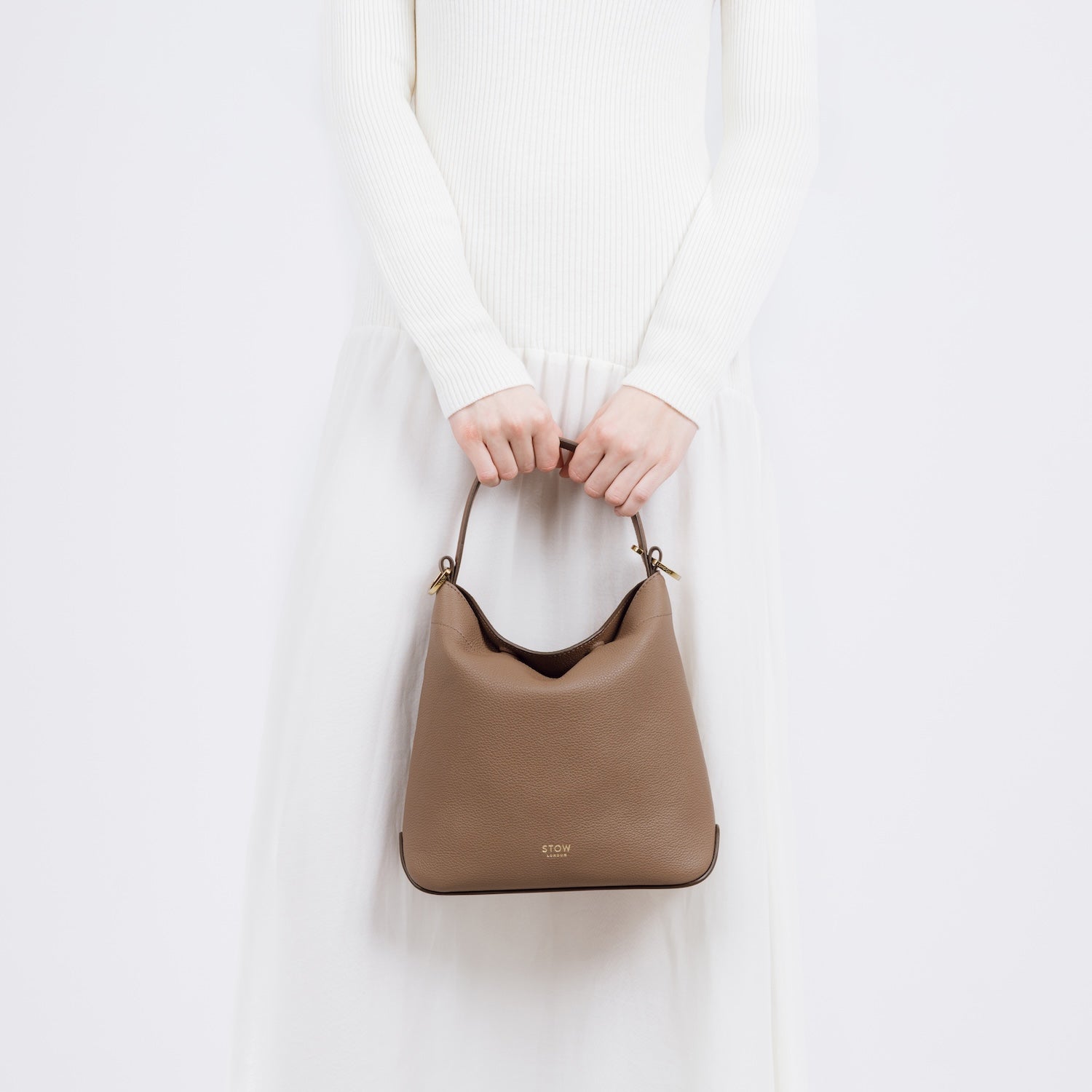 Model holding STOW London's Midi Curve in Mocha pebbled leather at her front featuring gold-coated stainless steel hardware