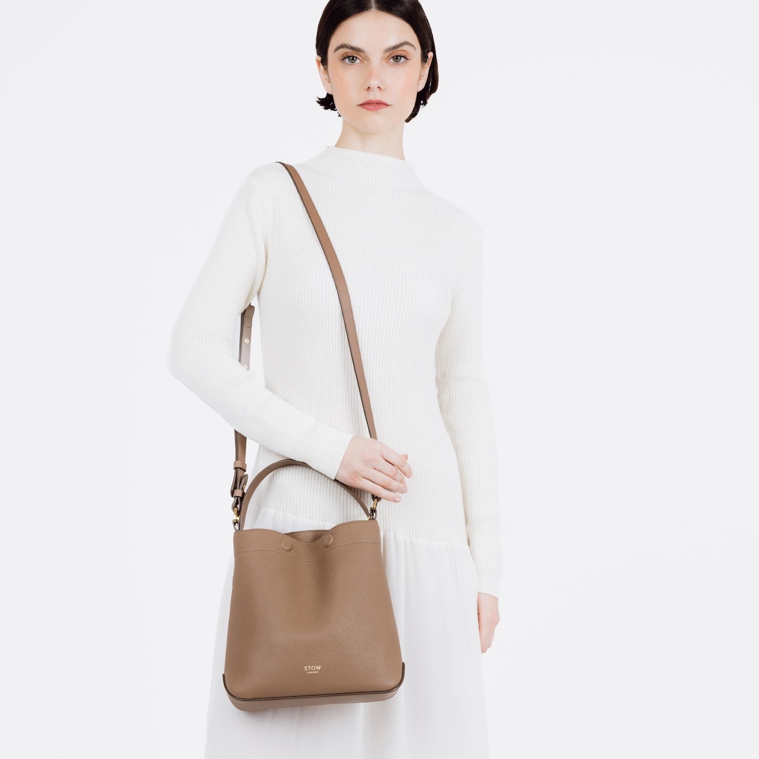 Model wearing STOW London's Midi Curve bag in Mocha pebbled and smooth leather on her shoulder featuring gold-coated stainless steel hardware on a white background