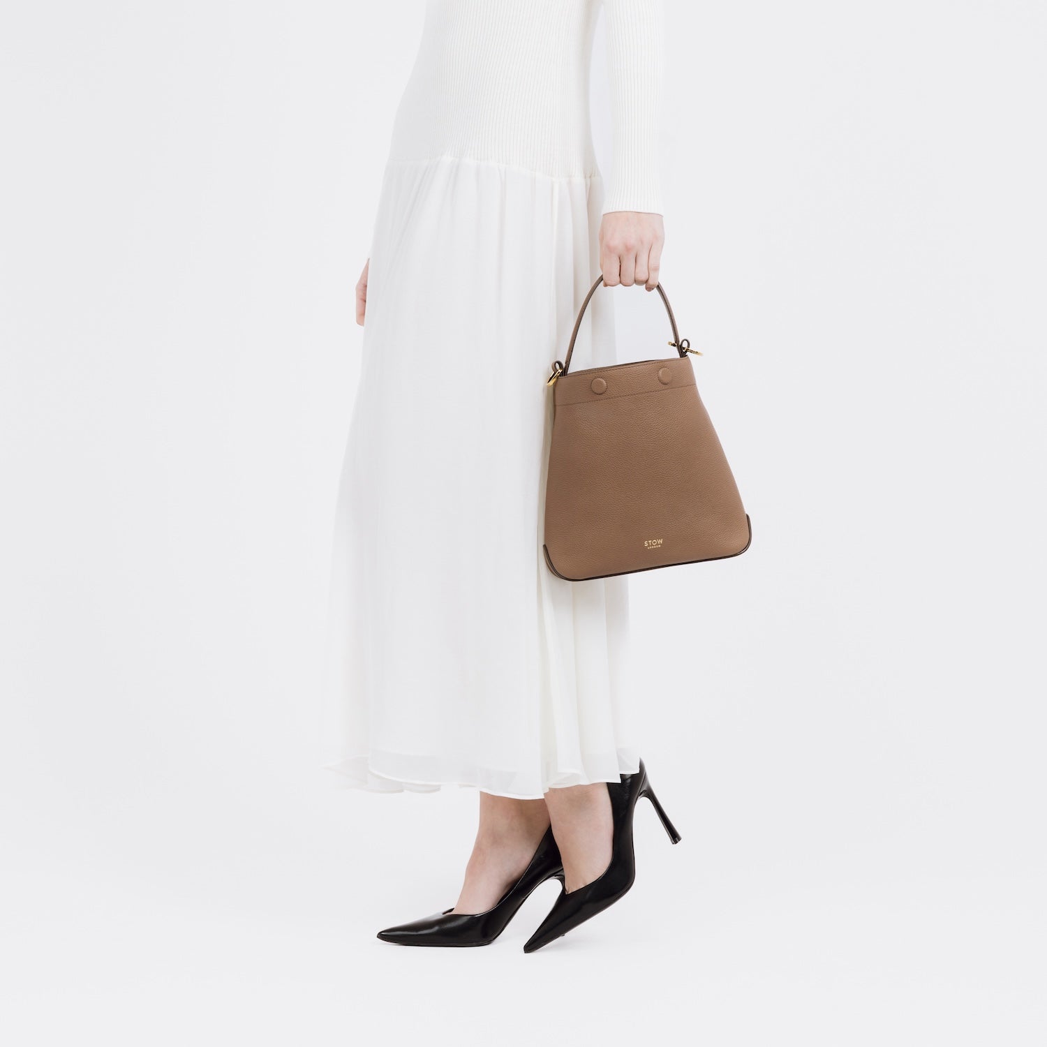 Model holding STOW London's Midi Curve in Mocha pebbled leather at her side featuring gold-coated stainless steel hardware