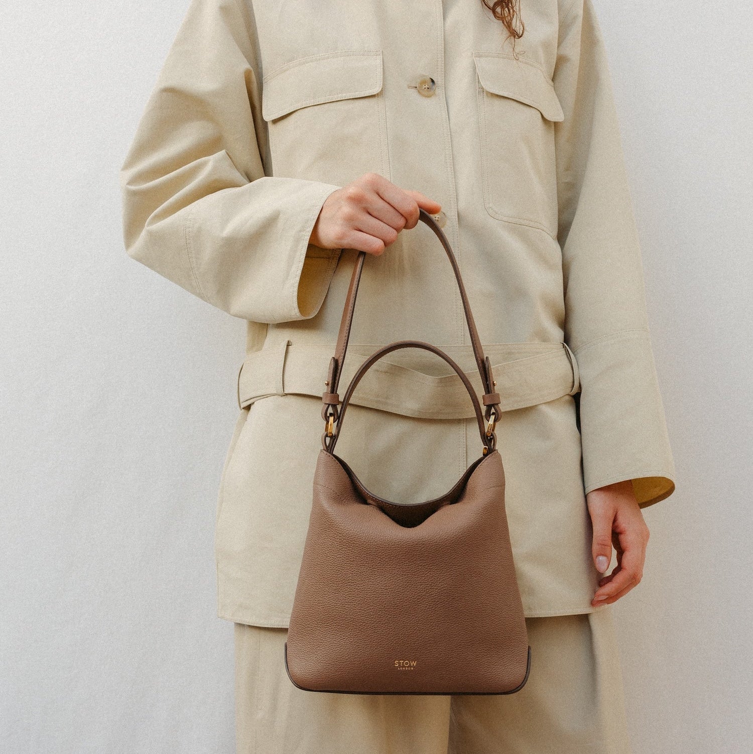 Model holding STOW London's Midi Curve in Mocha leather featuring the detachable shoulder strap and top handle with gold hardware and details