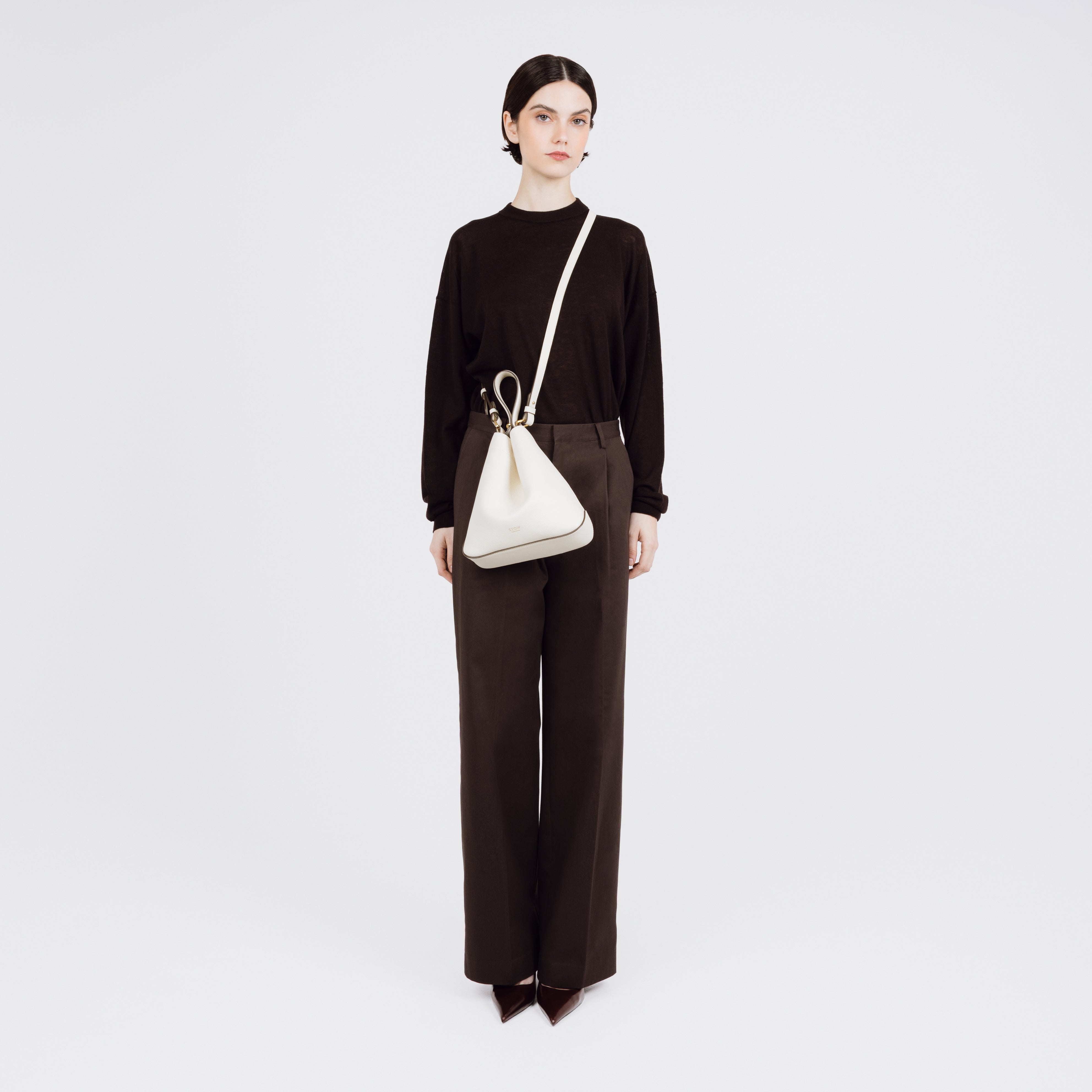 Model wearing STOW London's Midi Curve bag in Spring Moon pebbled and smooth leather crossbody styled in a triangular shape featuring gold-coated stainless steel hardware on a white background