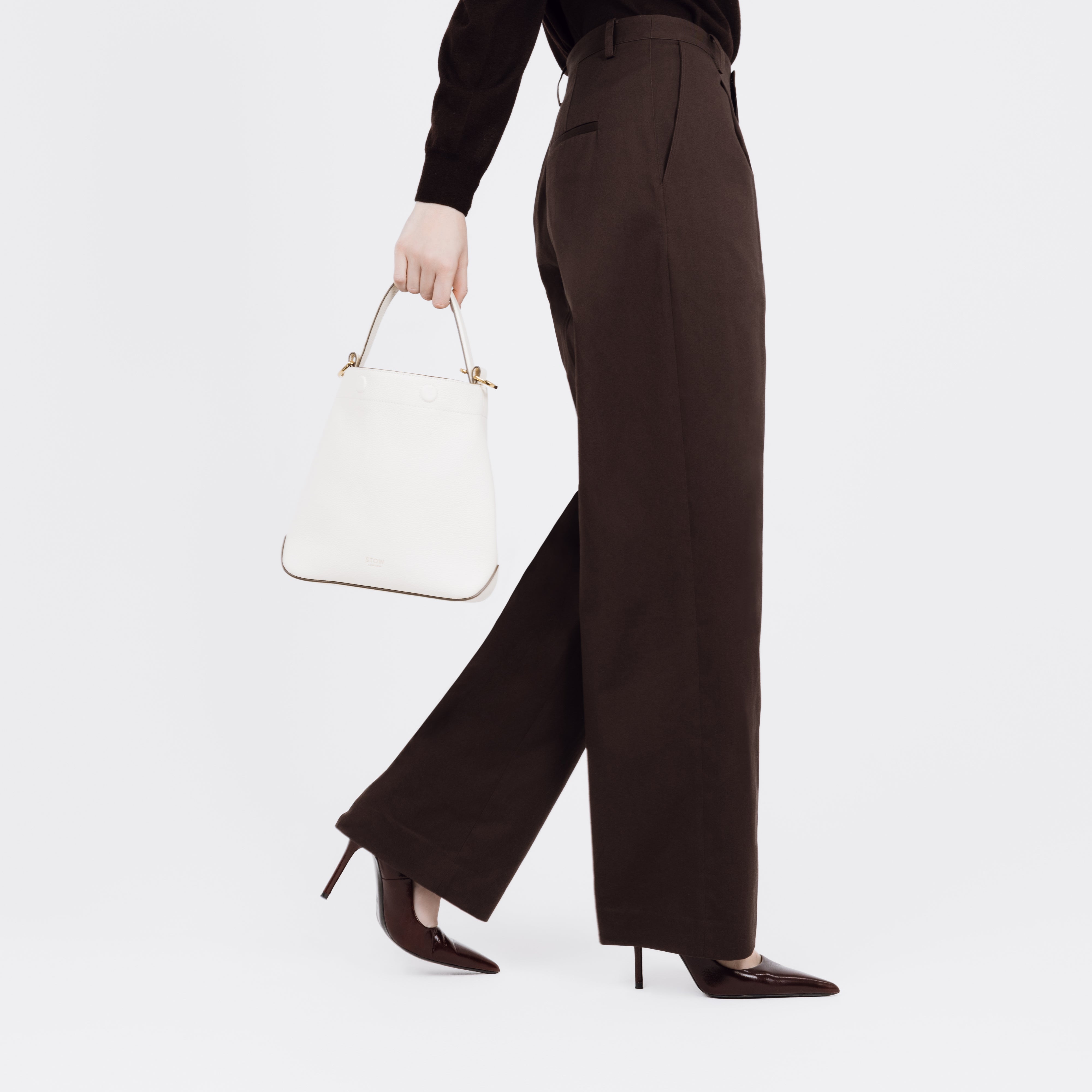 Model holding STOW London's Midi Curve bag in Spring Moon pebbled and smooth leather at her side styled as a bucket bag featuring gold-coated stainless steel hardware on a white background