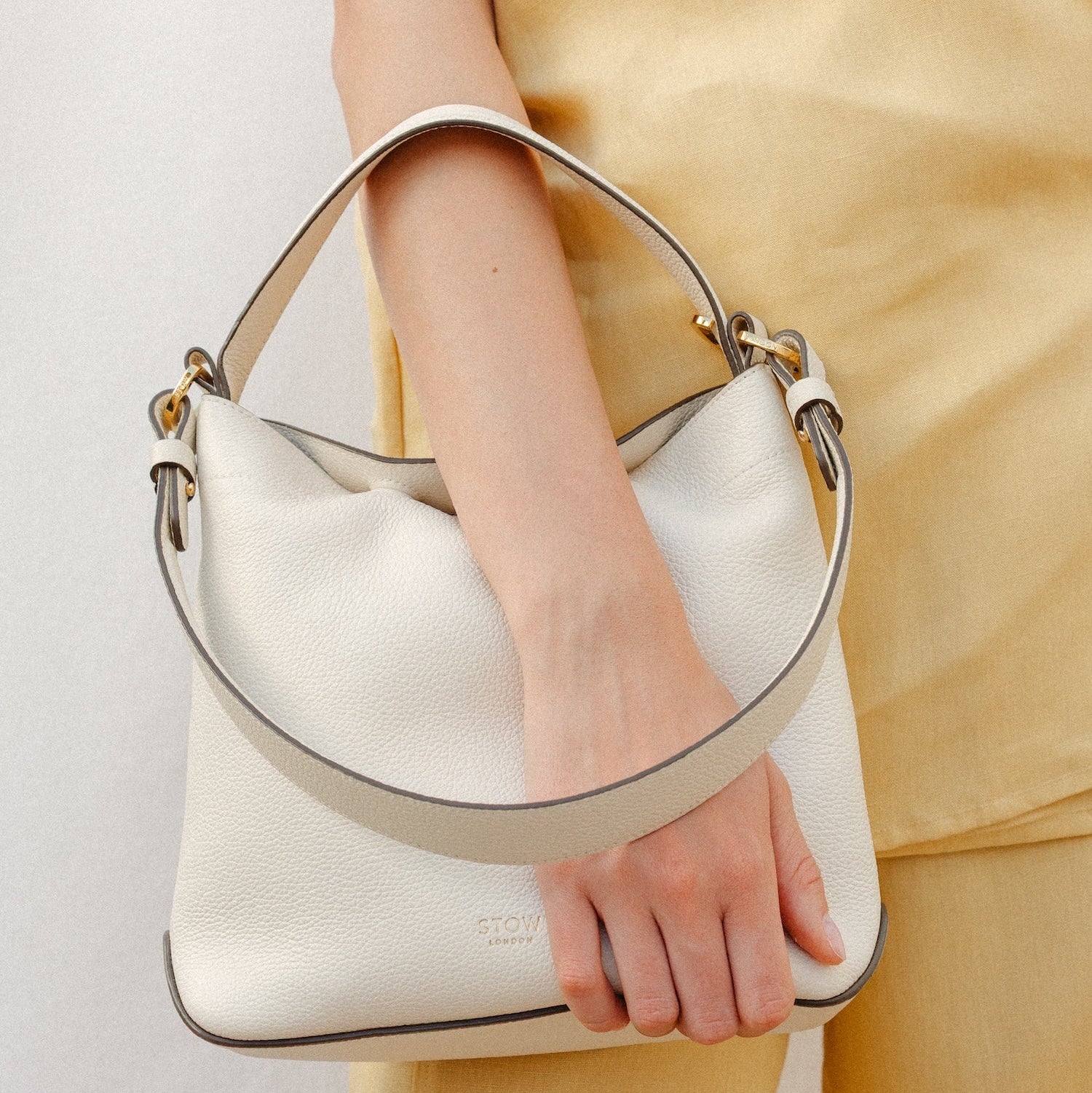 Model holding Model wearing STOW London's Midi Curve in Spring Moon leather featuring the detachable shoulder strap and top handle with gold hardware and details