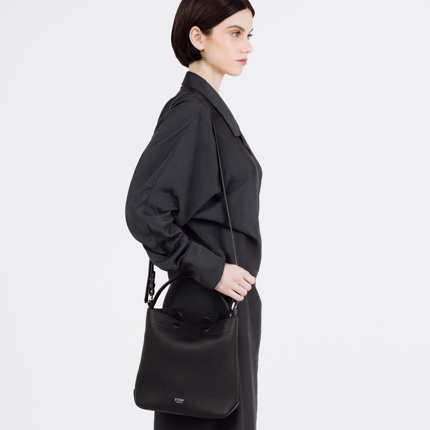 Model wearing STOW London's Midi Curve bag in Black pebbled and smooth leather on her shoulder featuring silver stainless steel hardware on a white background
