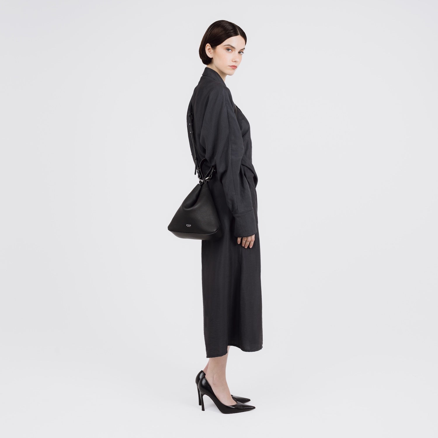 Model wearing STOW London's Midi Curve bag in Black pebbled and smooth leather crossbody in the soft triangular style featuring silver stainless steel hardware on a white background