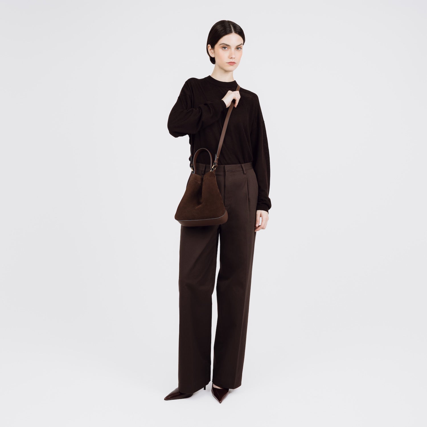 Model wearing STOW London's Midi Curve bag in Chocolate Suede leather crossbody styled in a triangular form featuring gold-coated stainless steel hardware on a white background