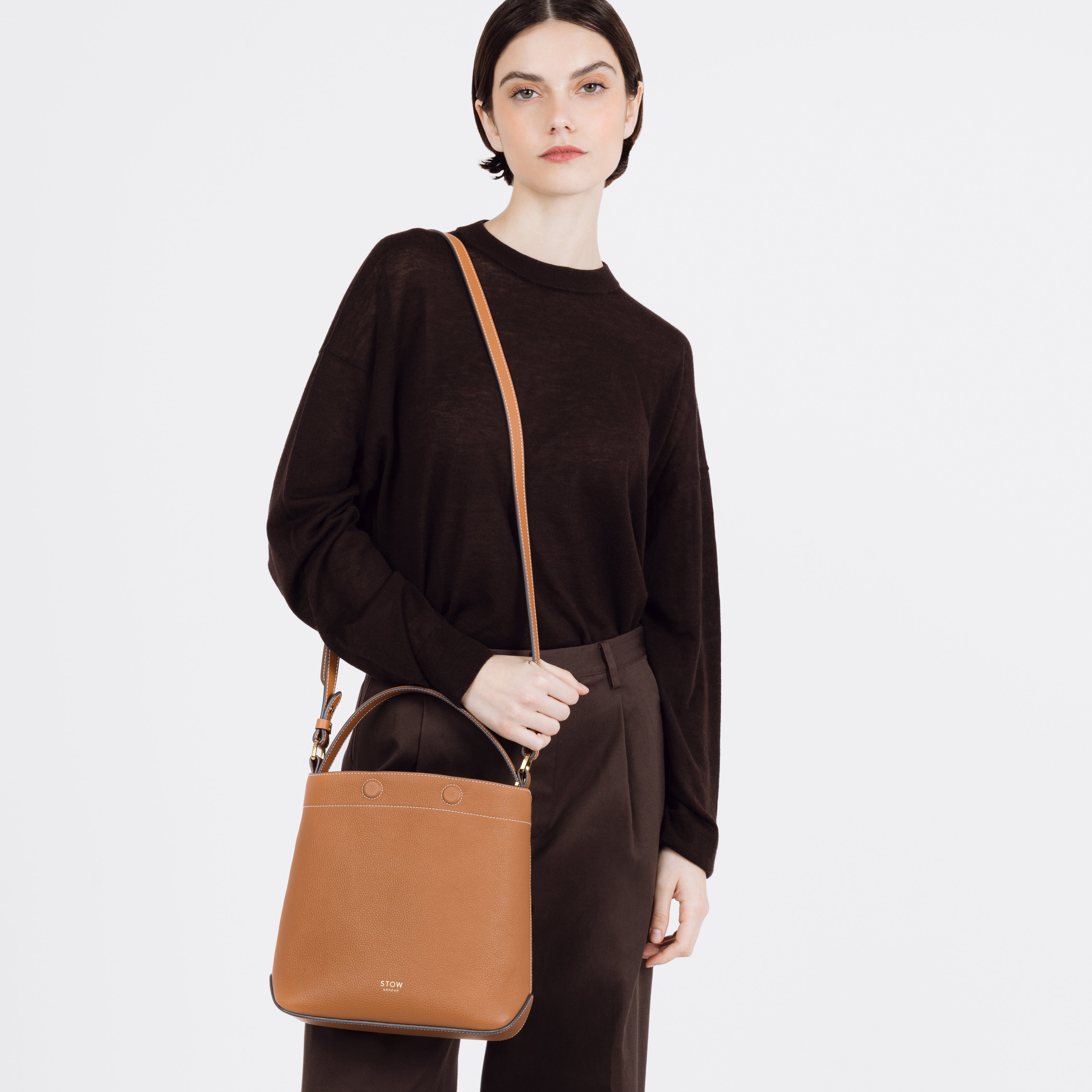 Model wearing STOW London's Midi Curve bag in Earth Tan pebbled and smooth leather on her shoulder shaped as a bucket bag featuring gold-coated stainless steel hardware on a white background