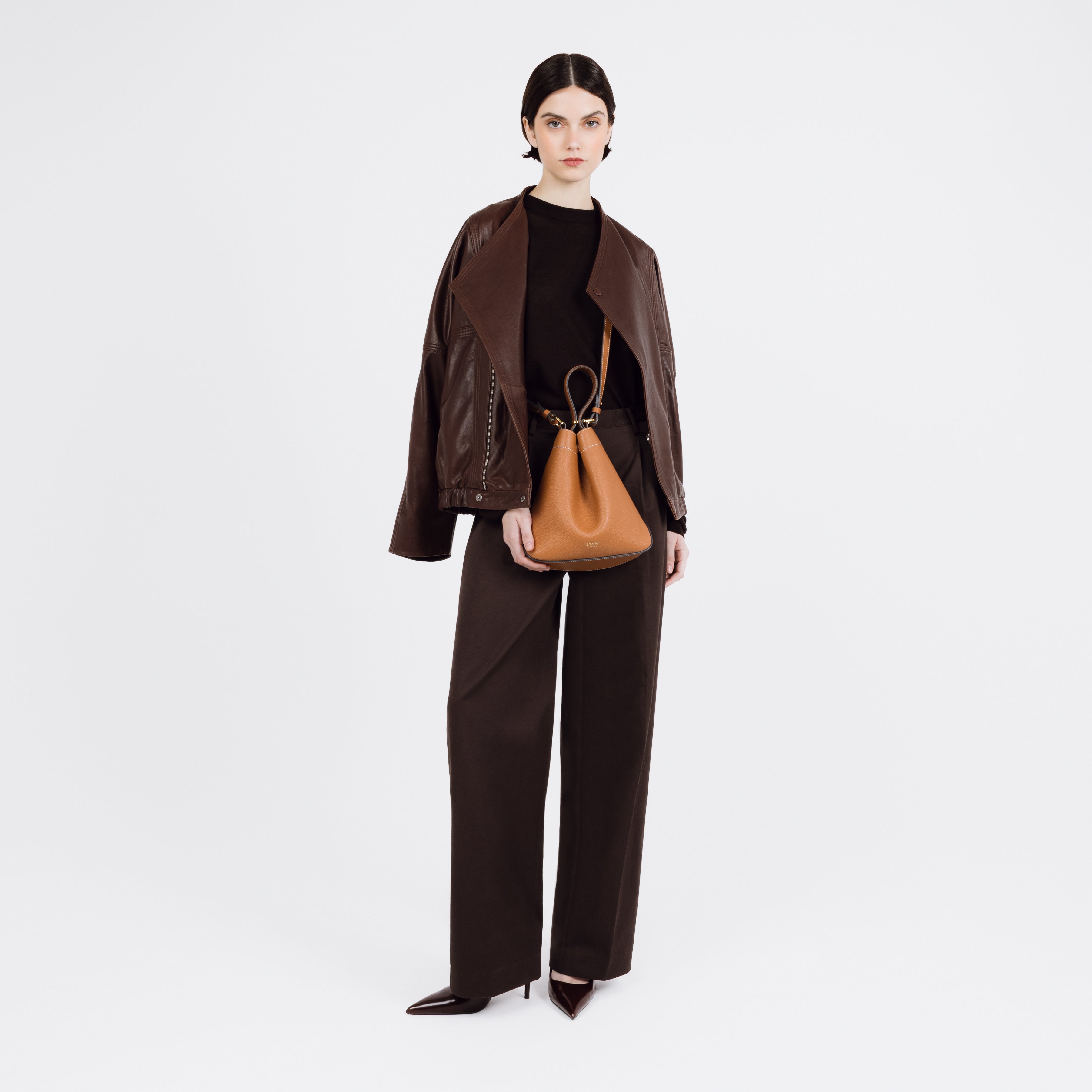 Model wearing STOW London's Midi Curve bag in Earth Tan pebbled and smooth leather crossbody styled in a triangular shape featuring gold-coated stainless steel hardware on a white background