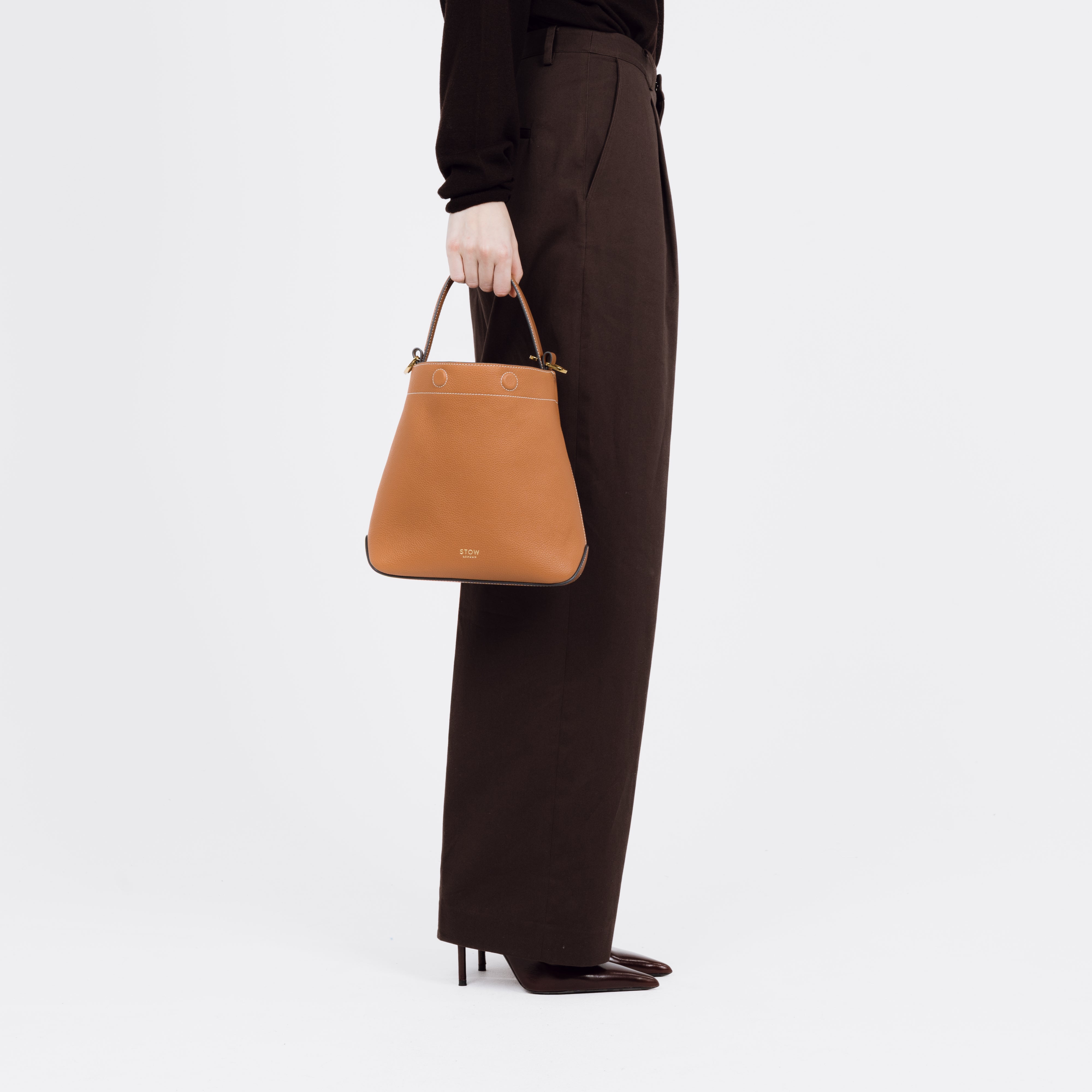 Model holding STOW London's Midi Curve bag in Earth Tan pebbled and smooth leather by top handle at her side shaped as a bucket bag featuring gold-coated stainless steel hardware on a white background