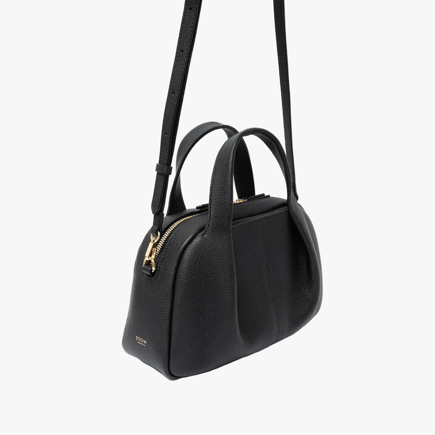 Off-axis view of STOW London's Mini Bowling bag in Black pebbled leather featuring a pleated front panel, detachable crossbody strap and gold-coated stainless steel hardware on a light grey background