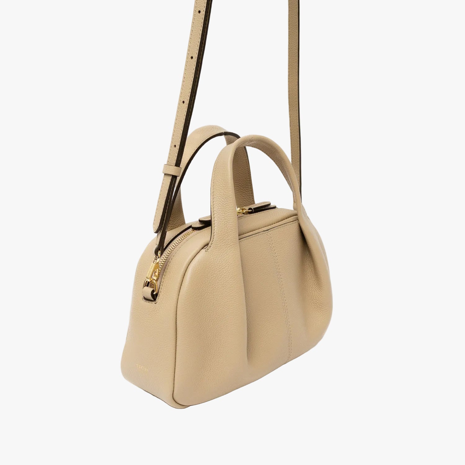 Off-axis view of STOW London's Mini Bowling bag in Camel pebbled leather featuring a pleated front panel, detachable crossbody strap and gold-coated stainless steel hardware on a light grey background