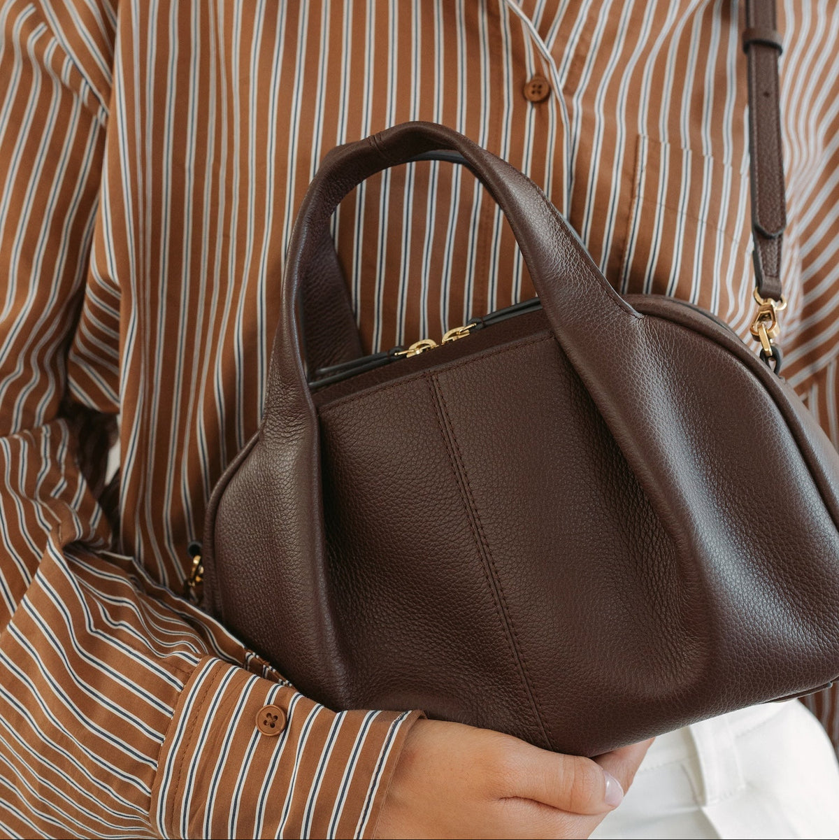 Model holding STOW London's Mini Bowling in Espresso pebbled leather featuring a pleated front panel and gold-coated stainless steel hardware