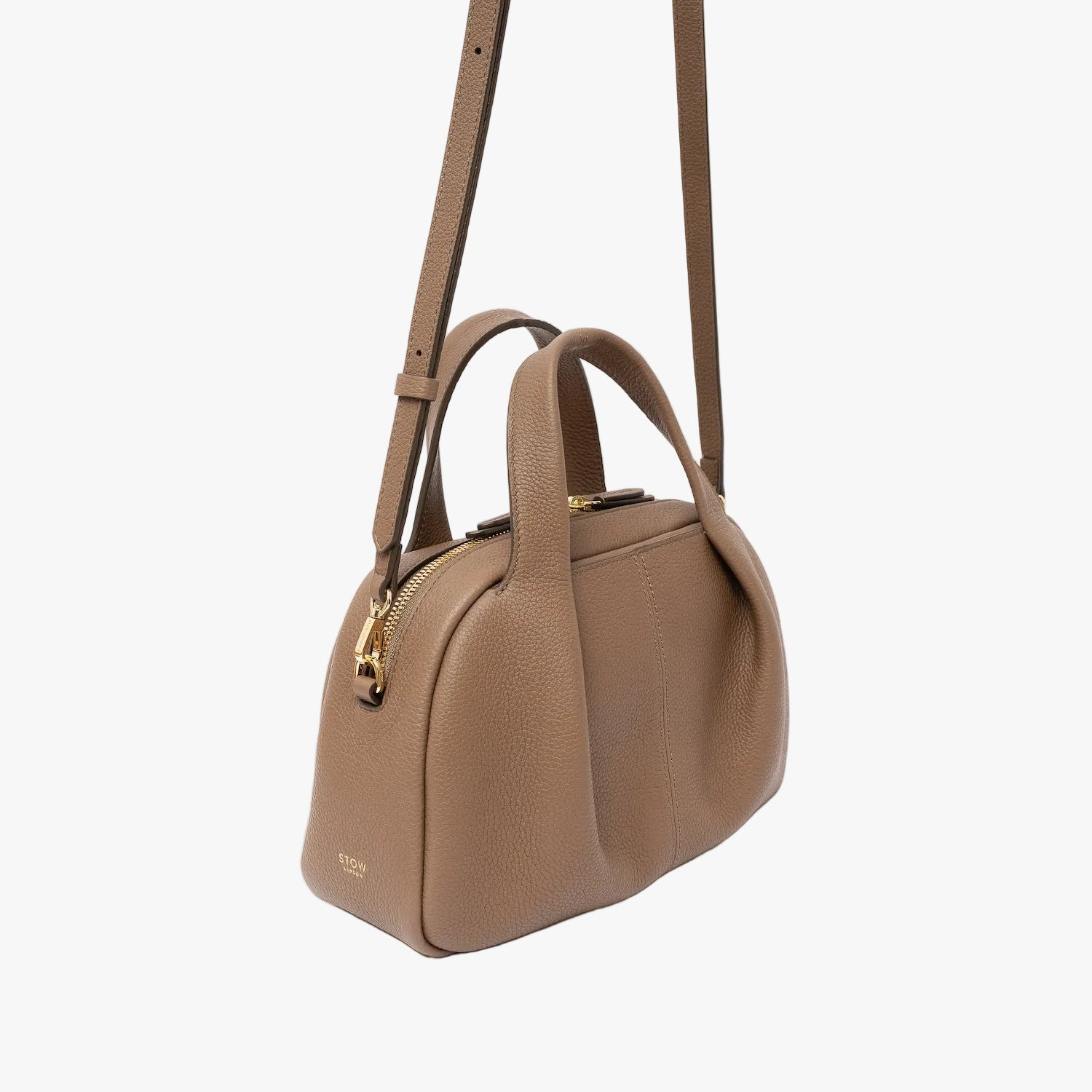 Off-axis view of STOW London's Mini Bowling bag in Mocha pebbled leather featuring a pleated front panel, the detachable crossbody strap and gold-coated stainless steel hardware on a light grey background