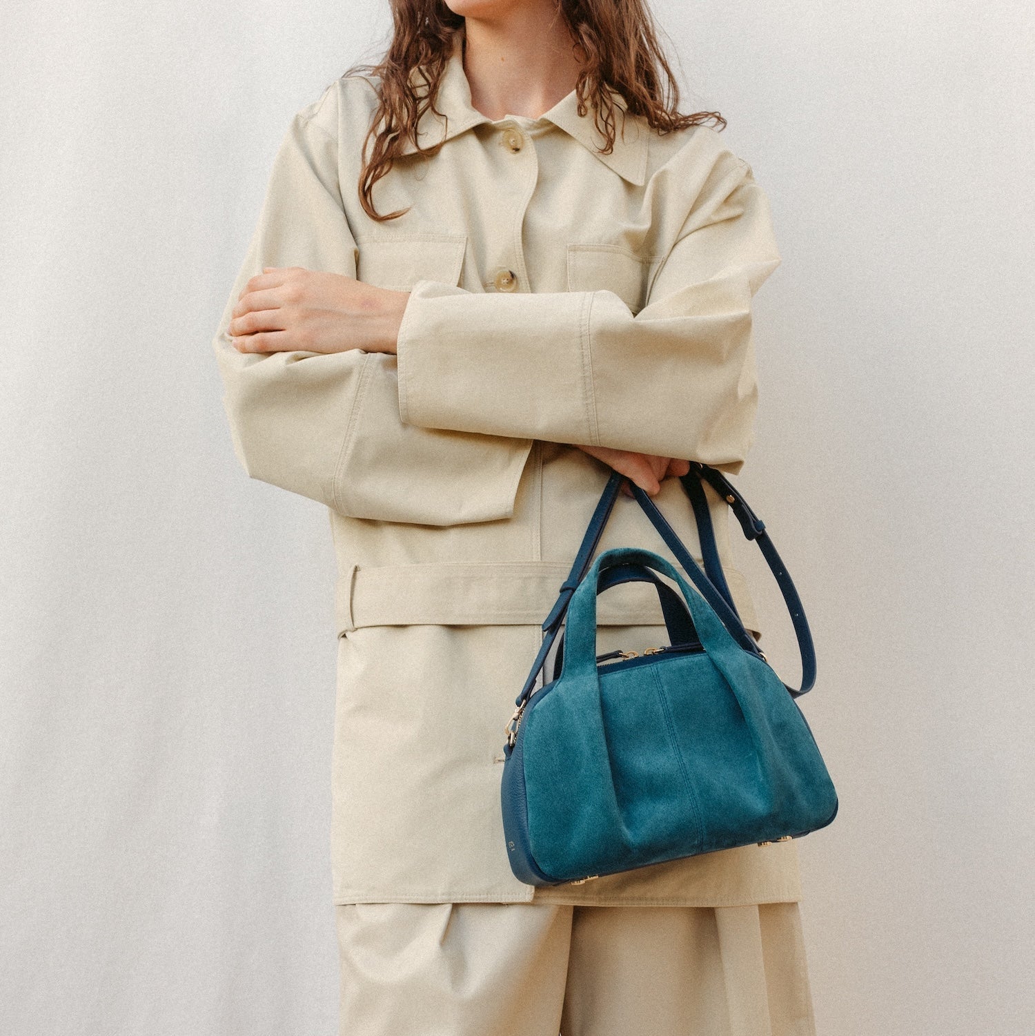 Model holding STOW London's Mini Bowling bag in Ocean Teal suede and smooth leather featuring a pleated front panel, detachable crossbody strap and gold-coated stainless steel hardware