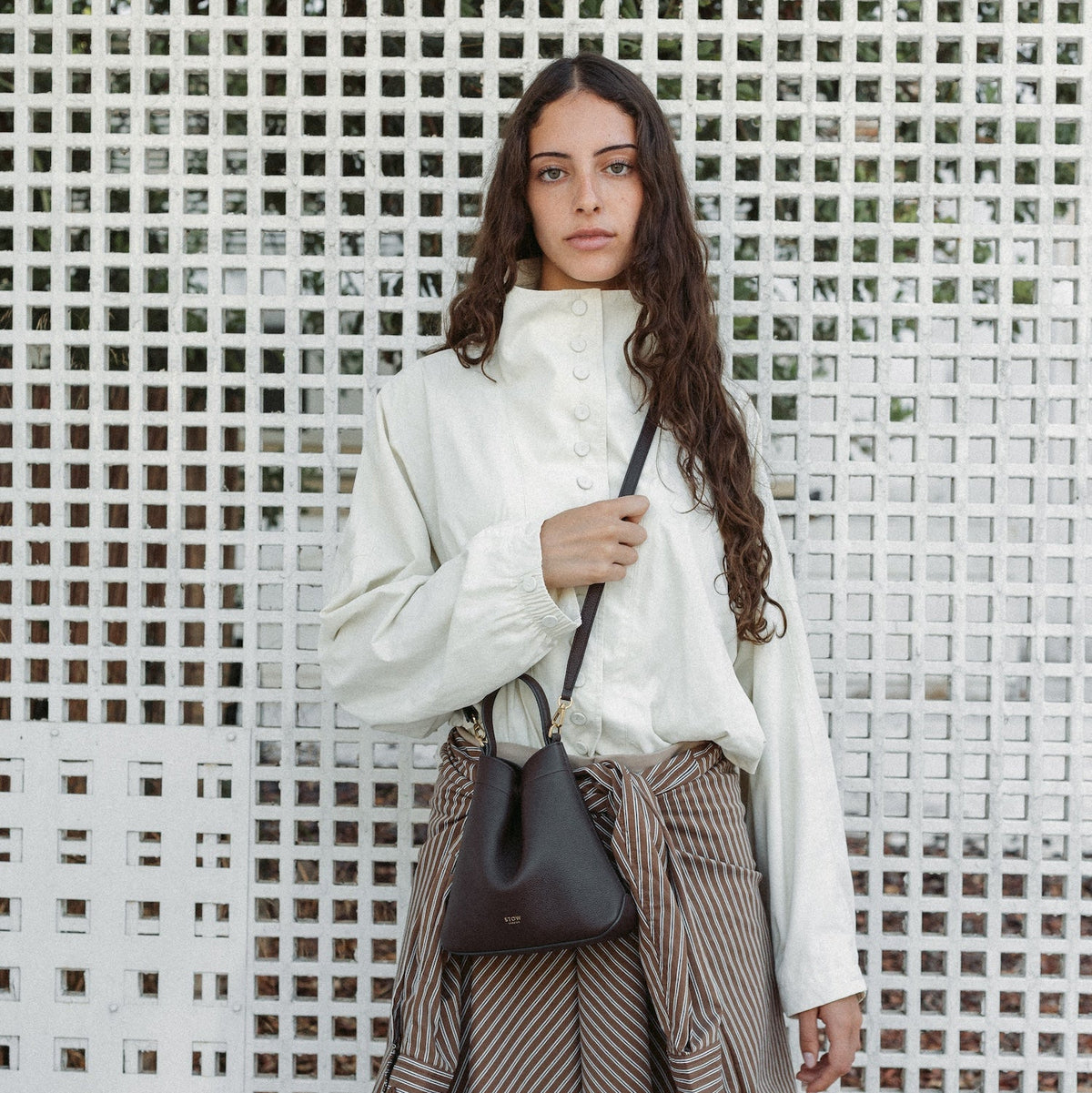 Model wearing STOW London's Mini Curve in Espresso pebbled leather crossbody featuring the detachable leather strap and gold-coated stainless steel hardware