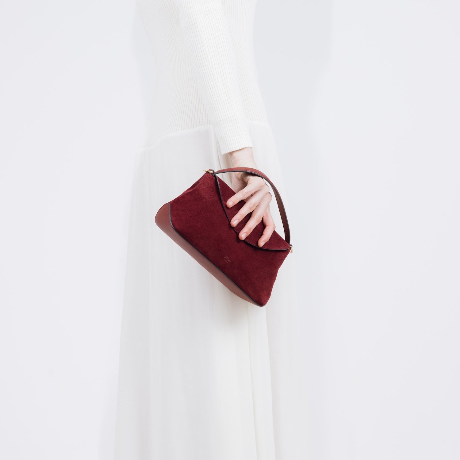 Model holding STOW London's Mini East West bag in Burgundy Suede leather as a clutch at her side featuring gold-coated stainless steel hardware on a white background
