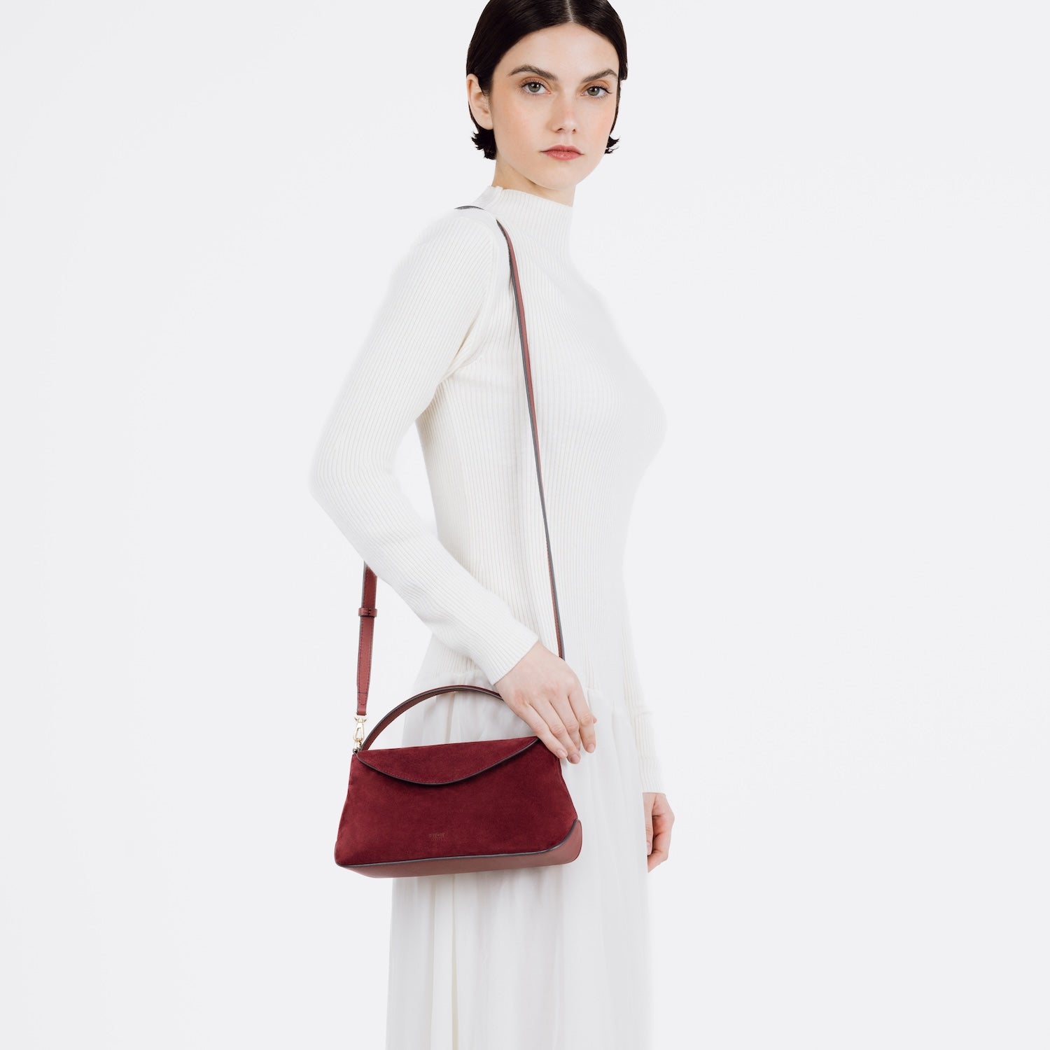 Model wearing STOW London's Mini East West bag in Burgundy Suede leather on her shoulder featuring gold-coated stainless steel hardware on a white background