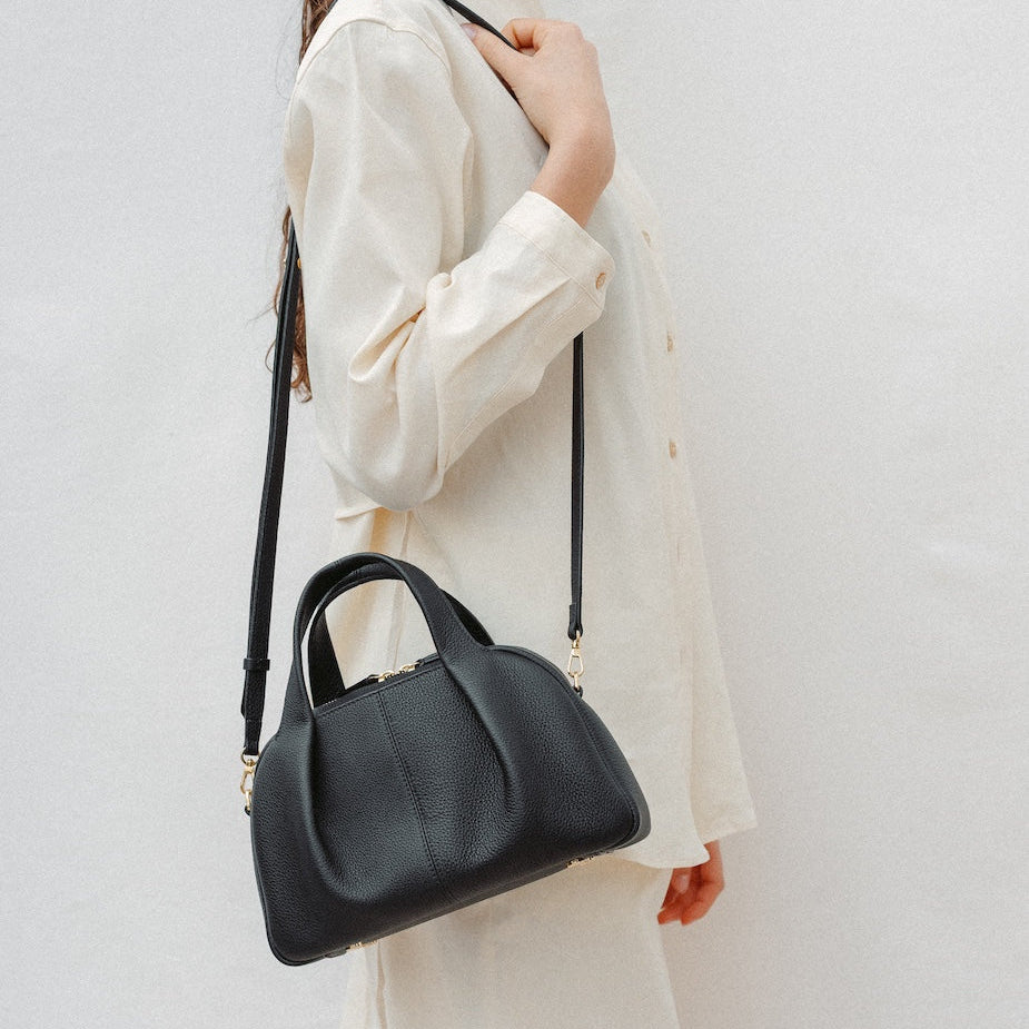 Model wearing STOW London's Mini Bowling in Black pebbled leather on her shoulder featuring a pleated front panel and gold-coated stainless steel hardware