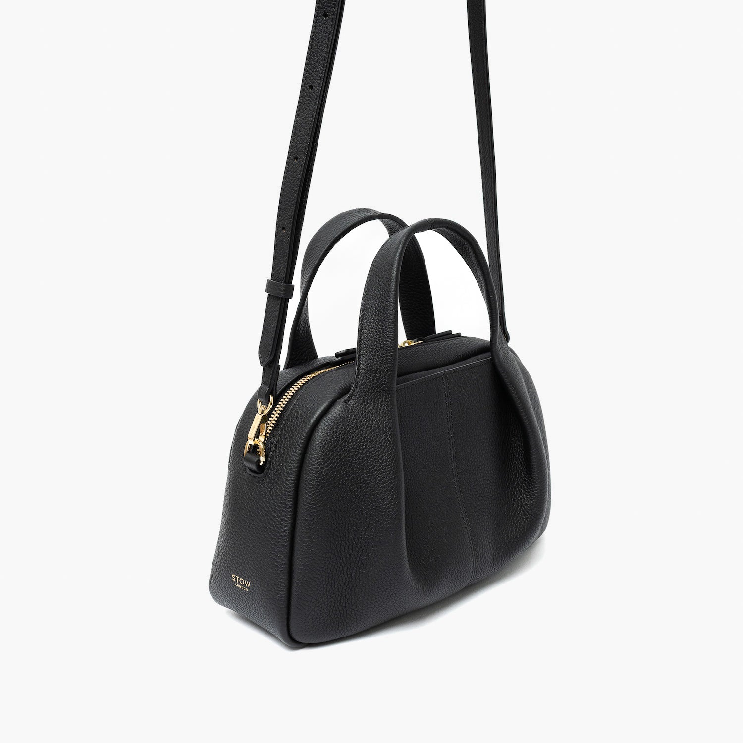 Off-axis view of STOW London's Mini Bowling bag in Black pebbled leather featuring a pleated front panel, detachable crossbody strap and gold-coated stainless steel hardware on a light grey background