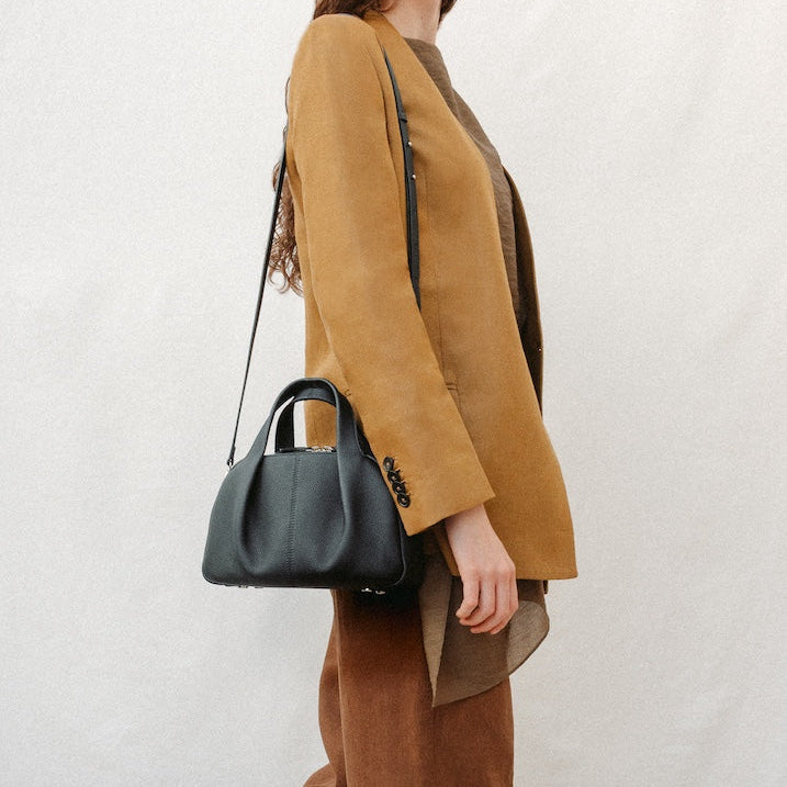 Model wearing STOW London's Mini Bowling in Black pebbled leather on her shoulder featuring a pleated front panel and silver stainless steel hardware