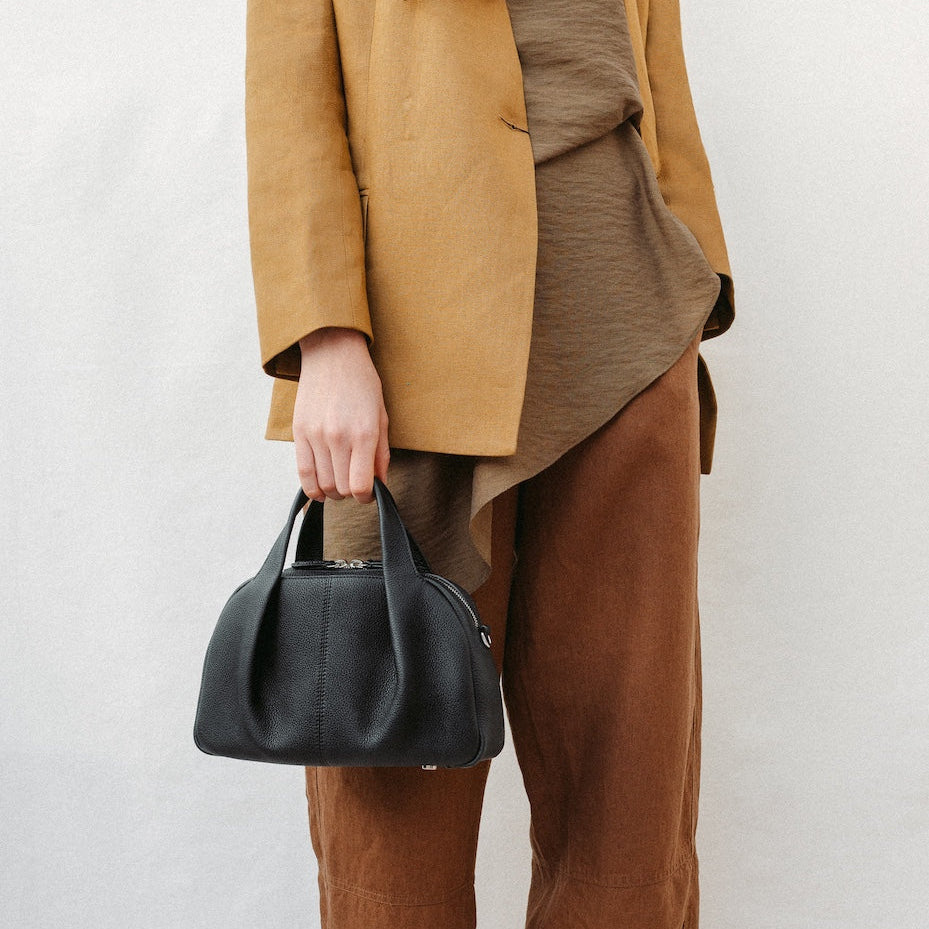 Model holding STOW London's Mini Bowling in Black pebbled leather at her side featuring a pleated front panel and silver stainless steel hardware