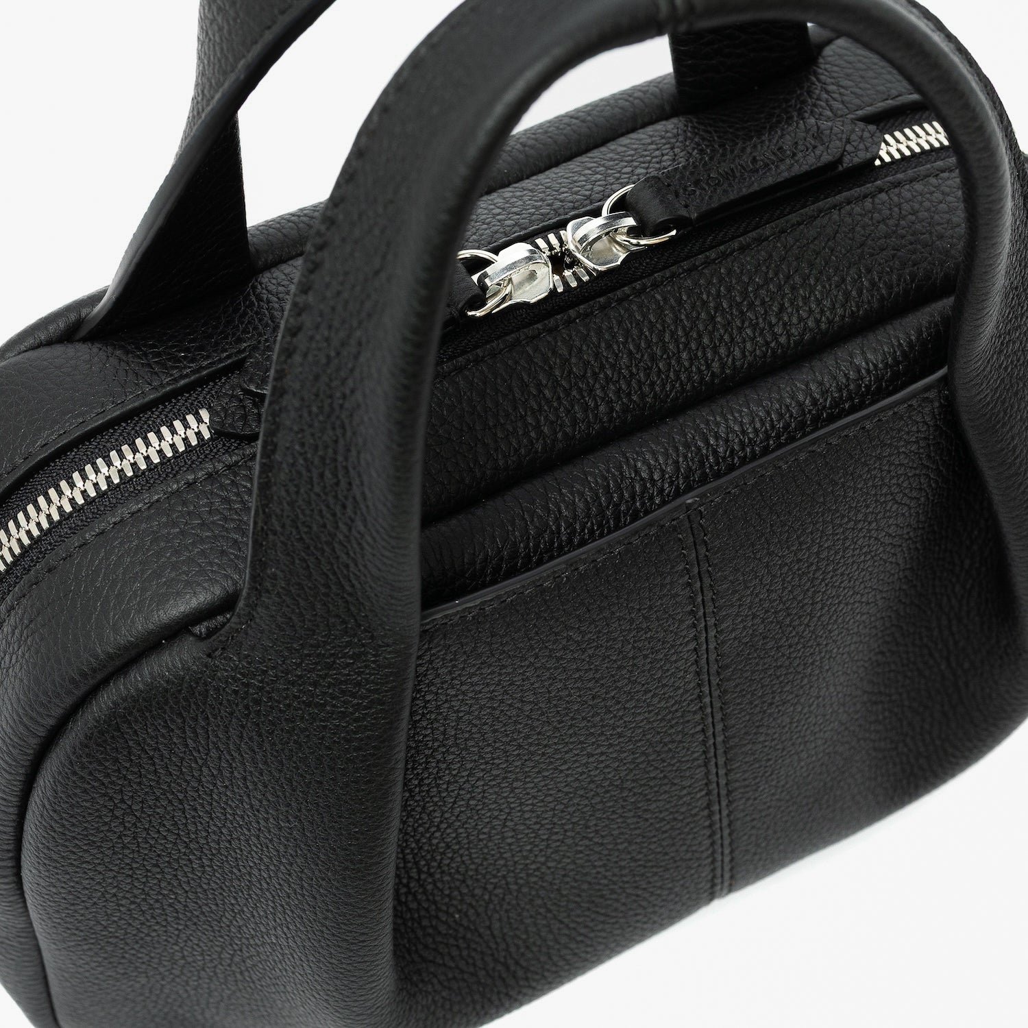 Close up of STOW London's Mini Bowling bag in Black pebbled leather featuring a pleated front panel with the external slip pocket in-between, zip closure, STOW London embossed pull tabs and silver stainless steel hardware on a light grey background