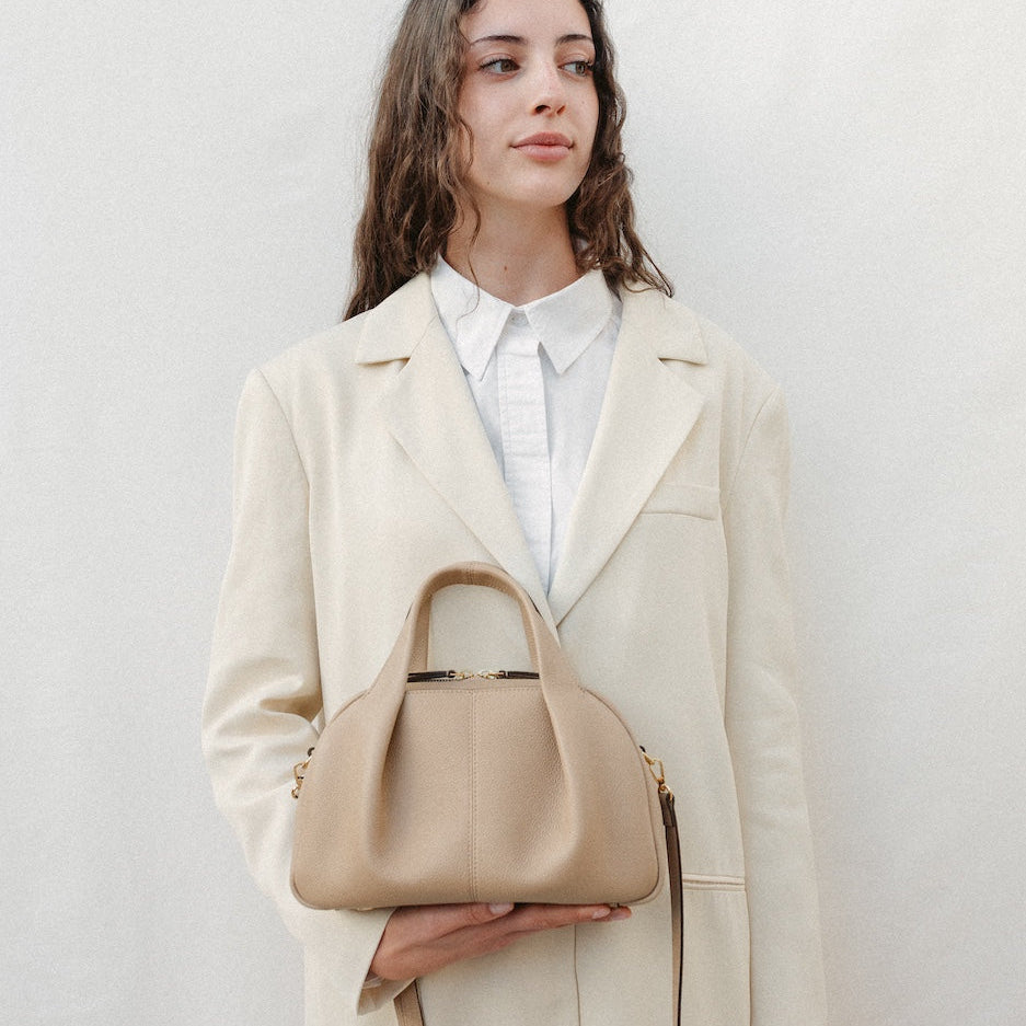 Model holding STOW London's Mini Bowling bag in Camel pebbled leather featuring a pleated front panel, detachable crossbody strap and gold-coated stainless steel hardware